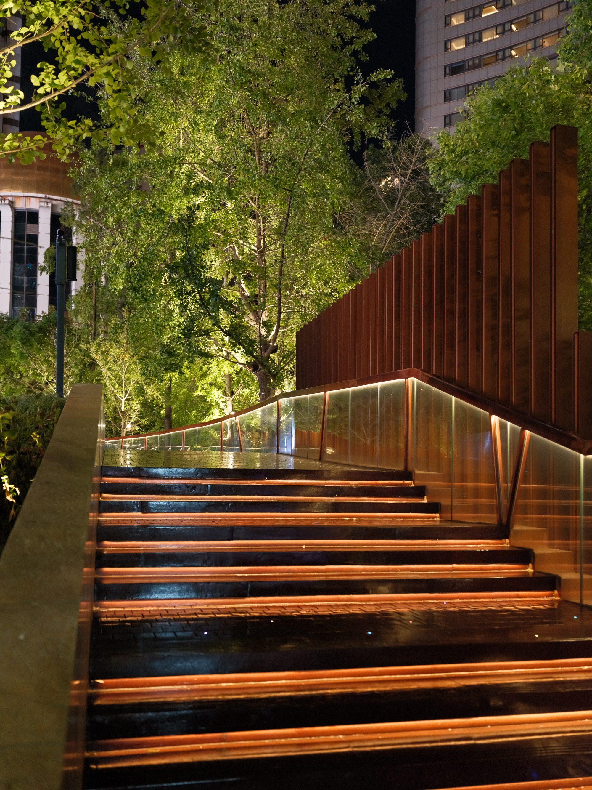 Night view of a lit stair with copper-edged treads, glass balustrade and weathering steel fins beneath illuminated trees