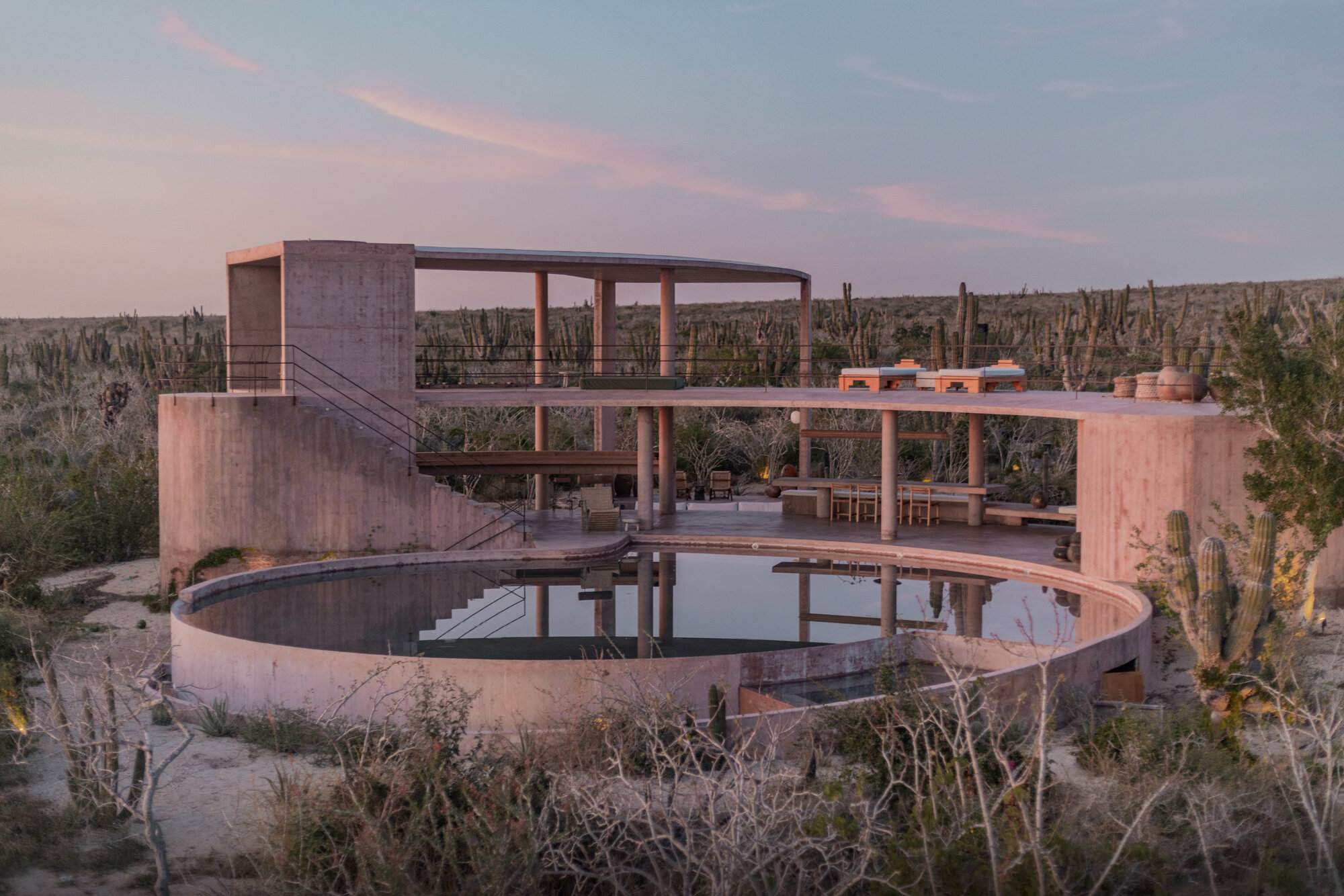 Circular pool and raised concrete pavilion with slender columns among tall cacti under a pink dusk sky