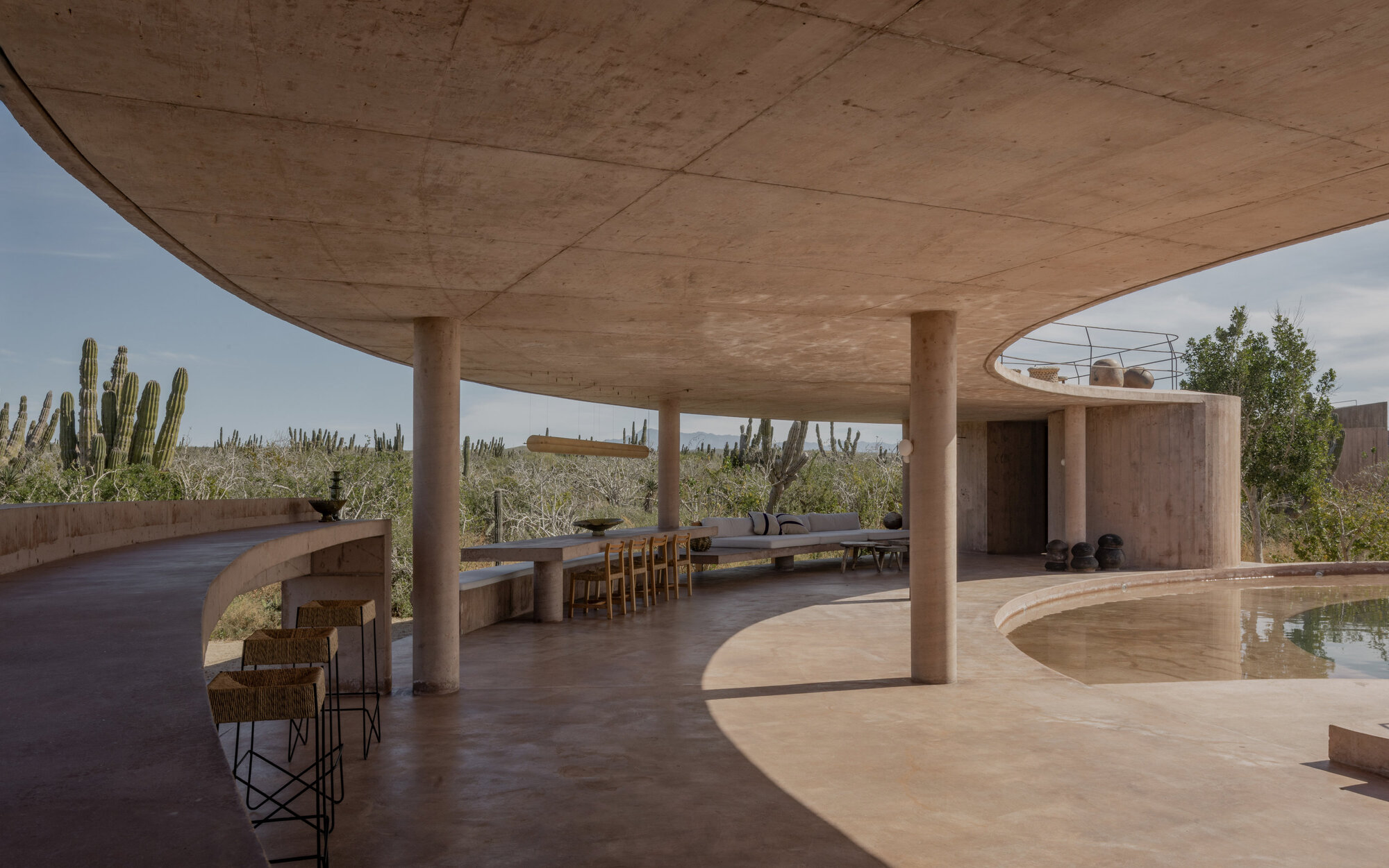 Curved concrete canopy over a long dining table and woven bar stools, with cacti beyond in soft daylight