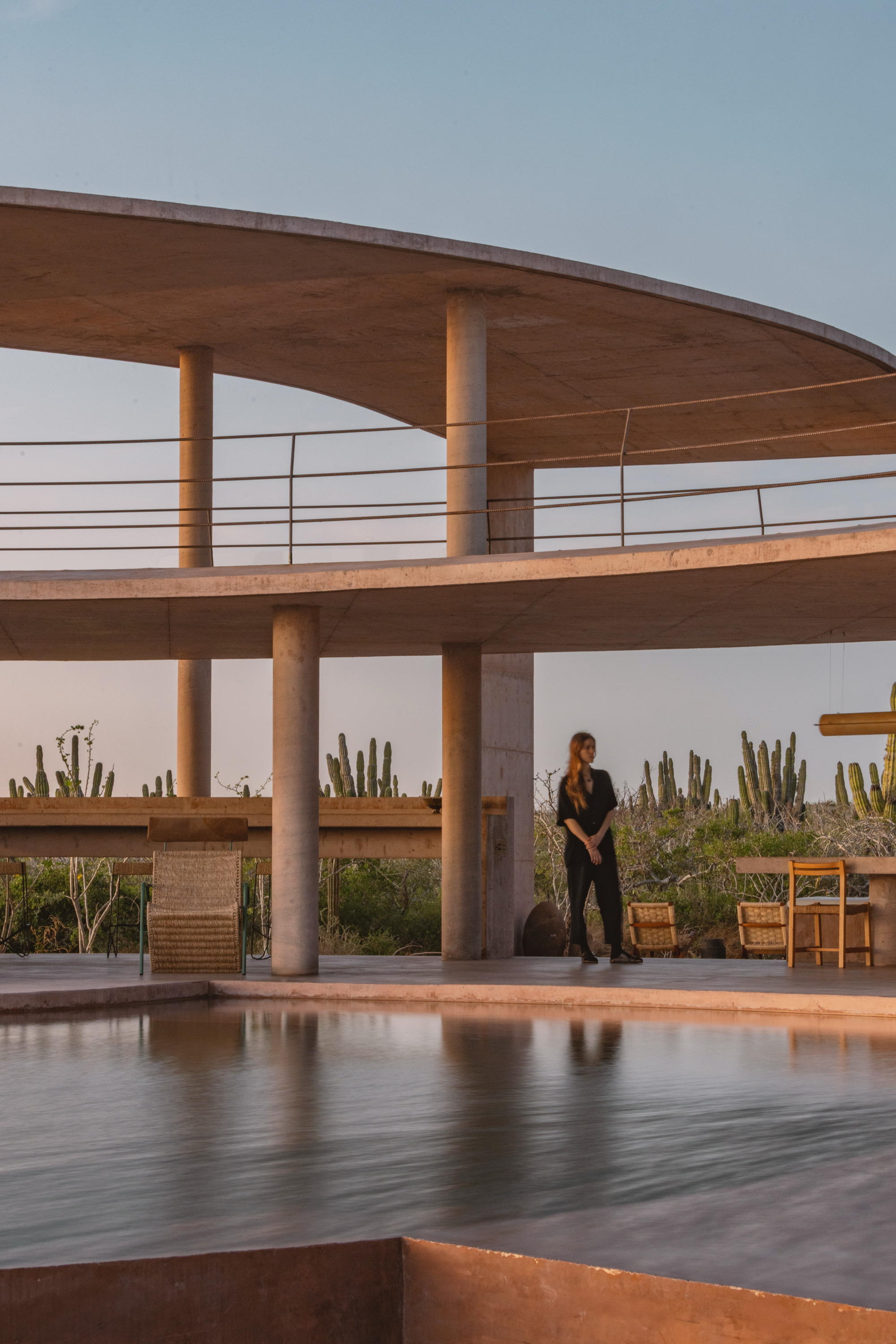 Woman standing between round concrete columns beside the pool, with stacked curved slabs and cacti behind at sunset