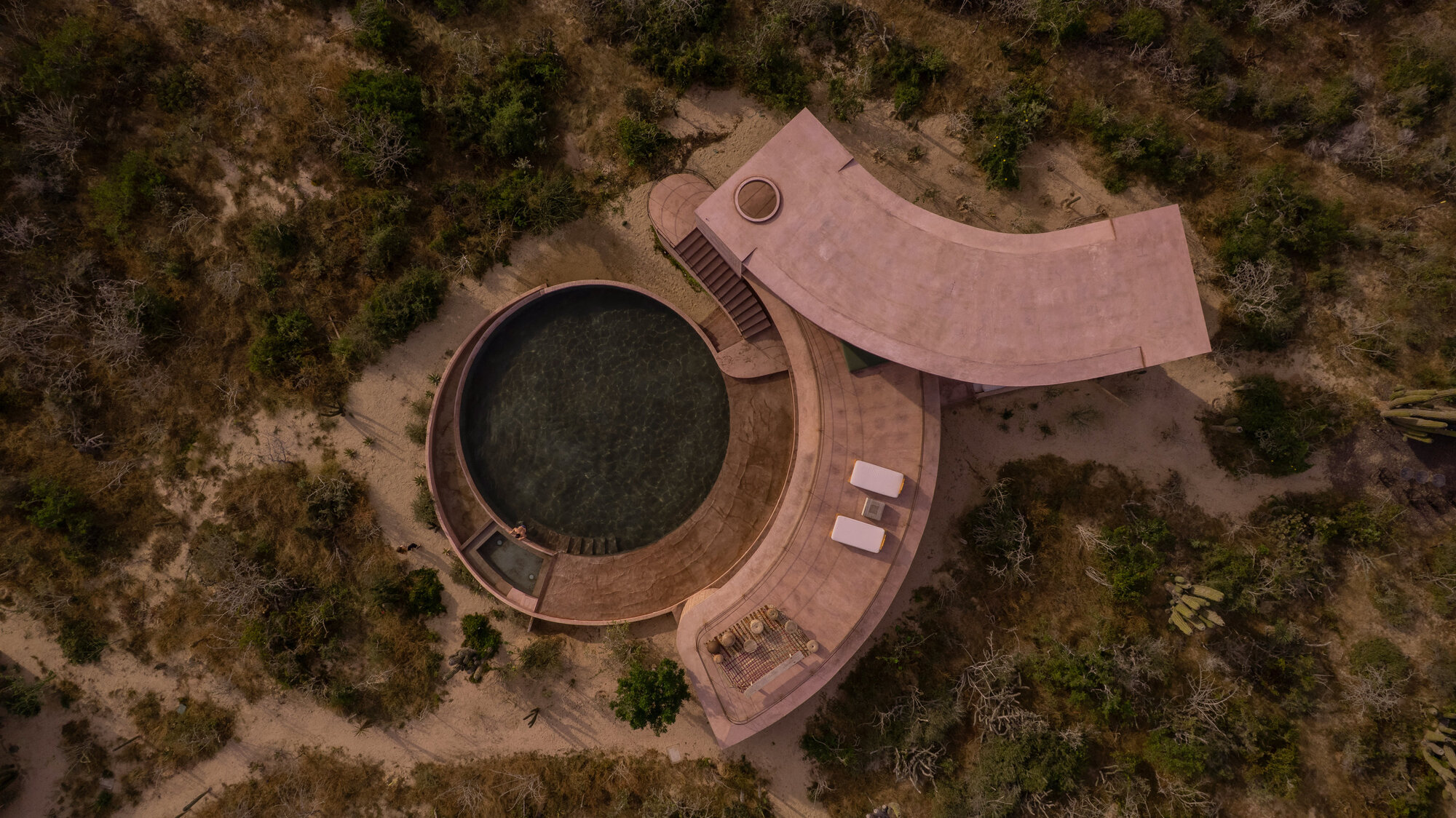 Drone view of the round pool and curved pink roof surrounded by desert scrub and sandy paths