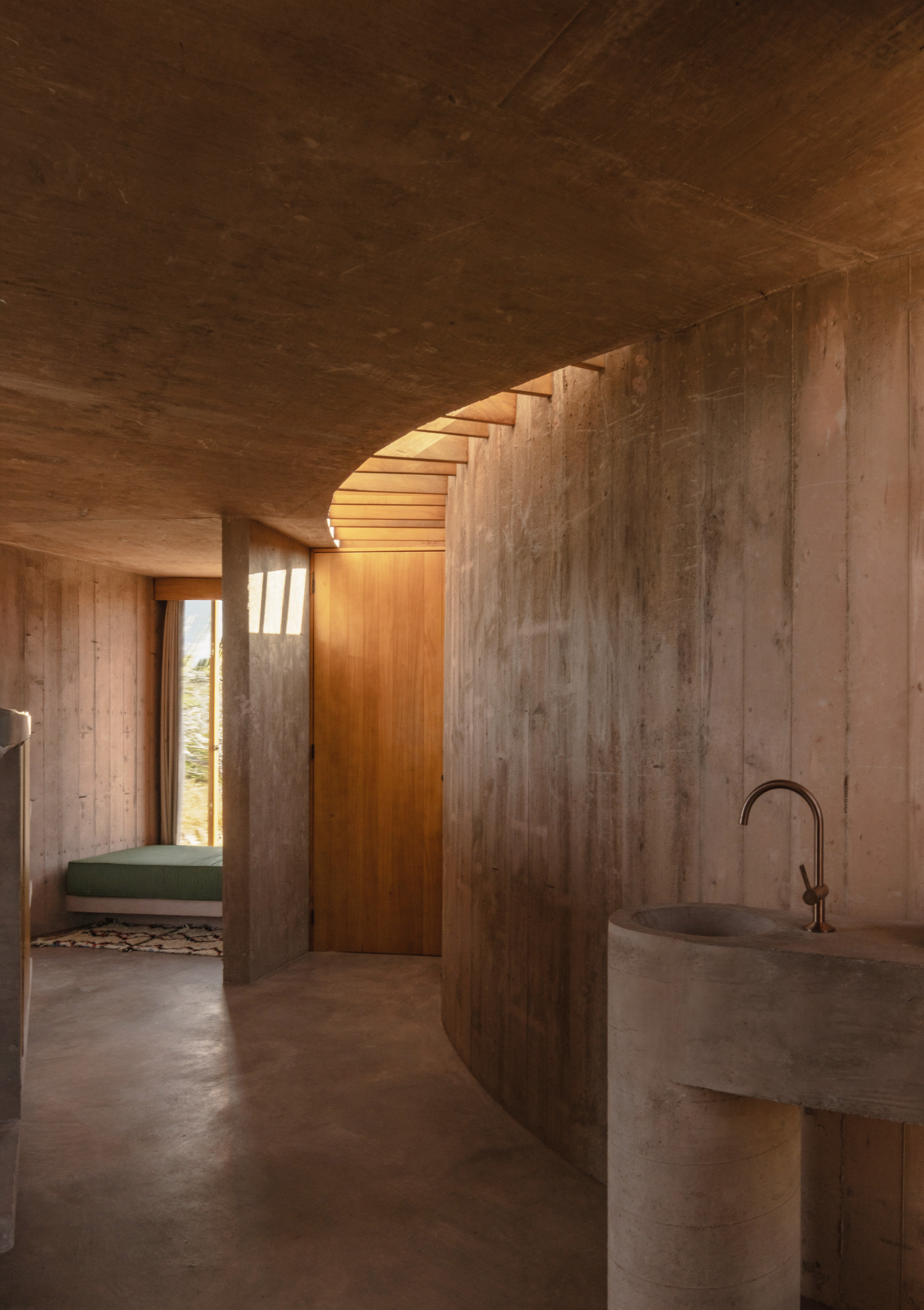 Curved board-formed concrete wall with a round concrete basin and brass tap, lit by warm light from a timber-slatted skylight