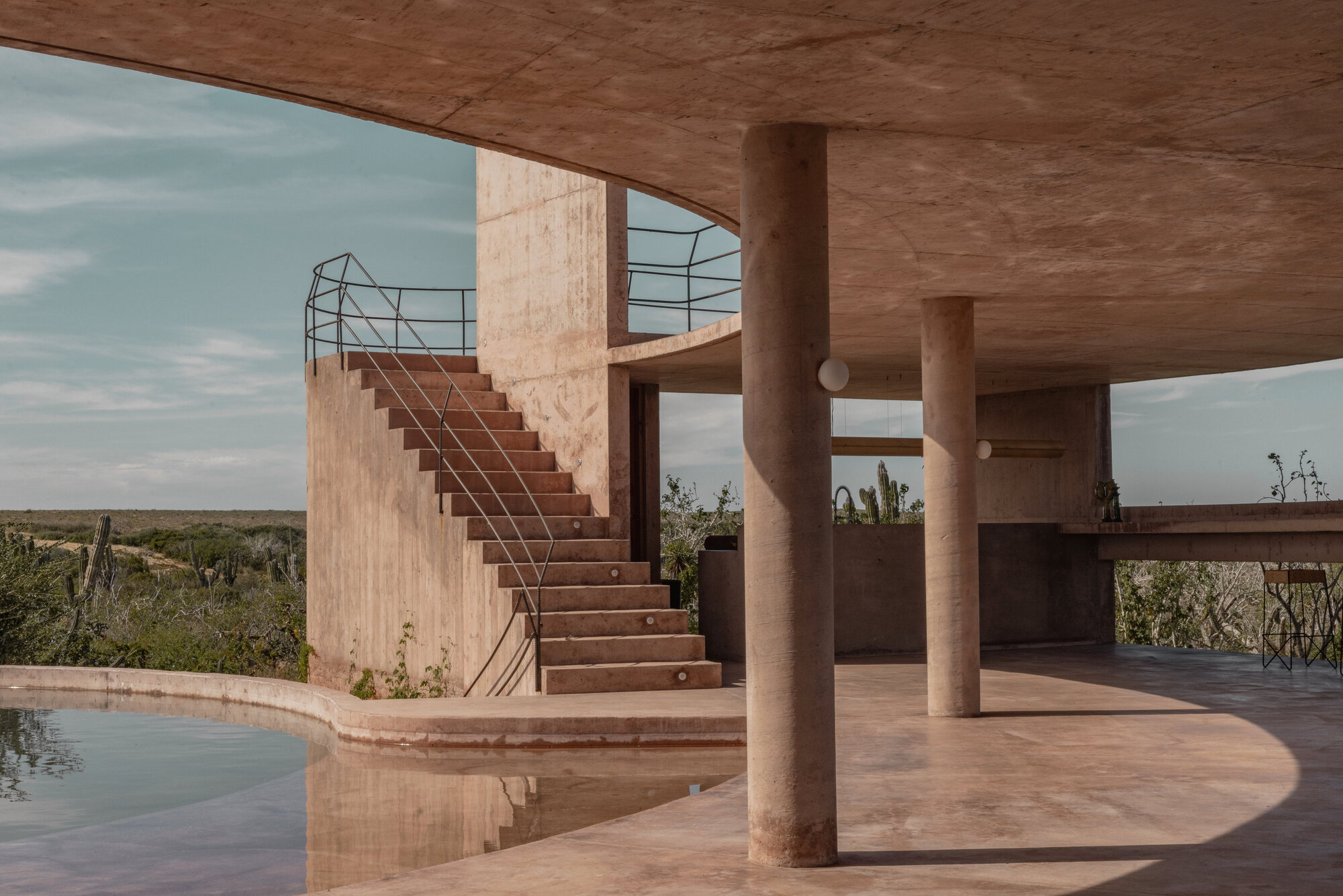 Concrete staircase with metal railings rising beside the pool, framed by round columns under a blue sky with thin clouds