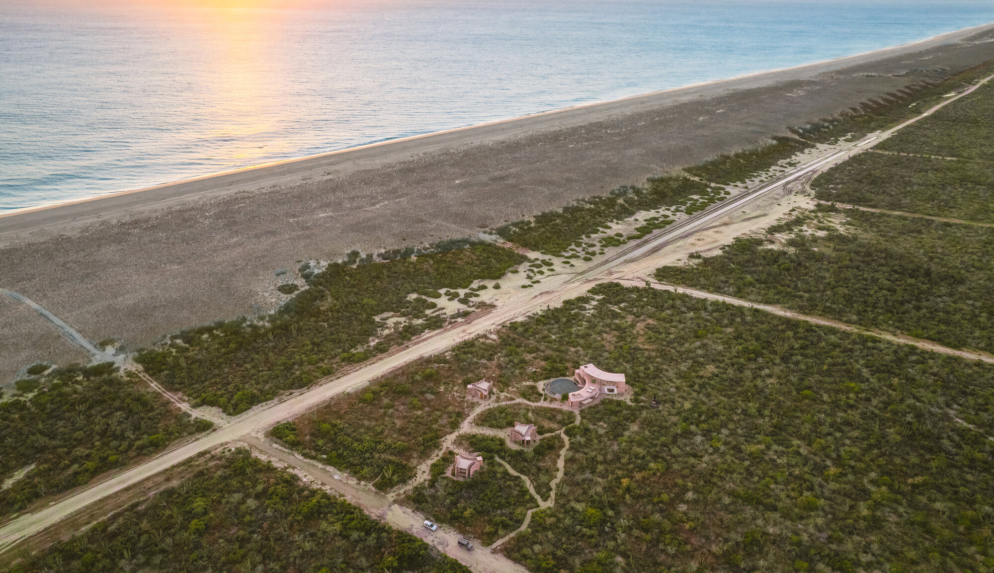 Aerial view of small pink-toned cabins and a round pool among scrub, beside a dirt road and long beach at sunset