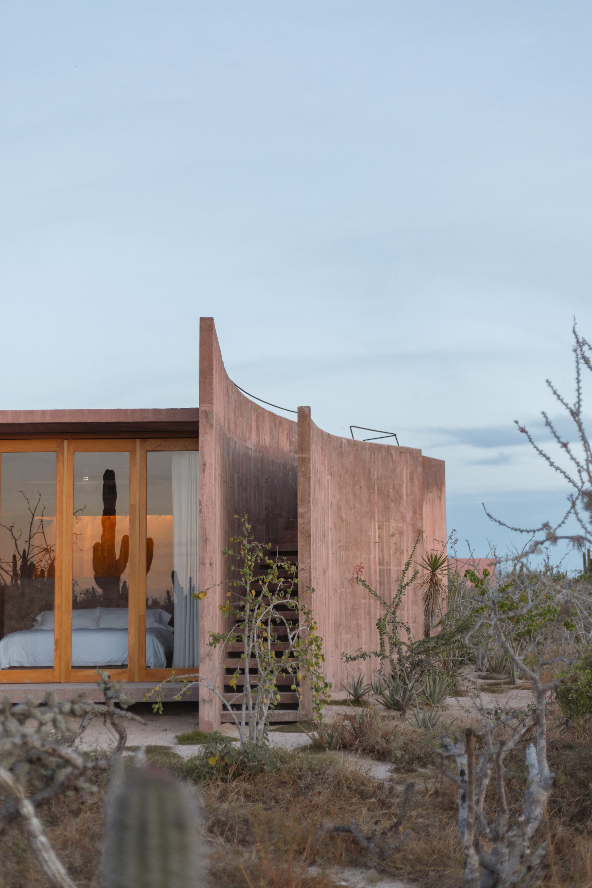 Glazed timber bedroom front beside a curved concrete wall and stair, surrounded by desert shrubs under a hazy evening sky