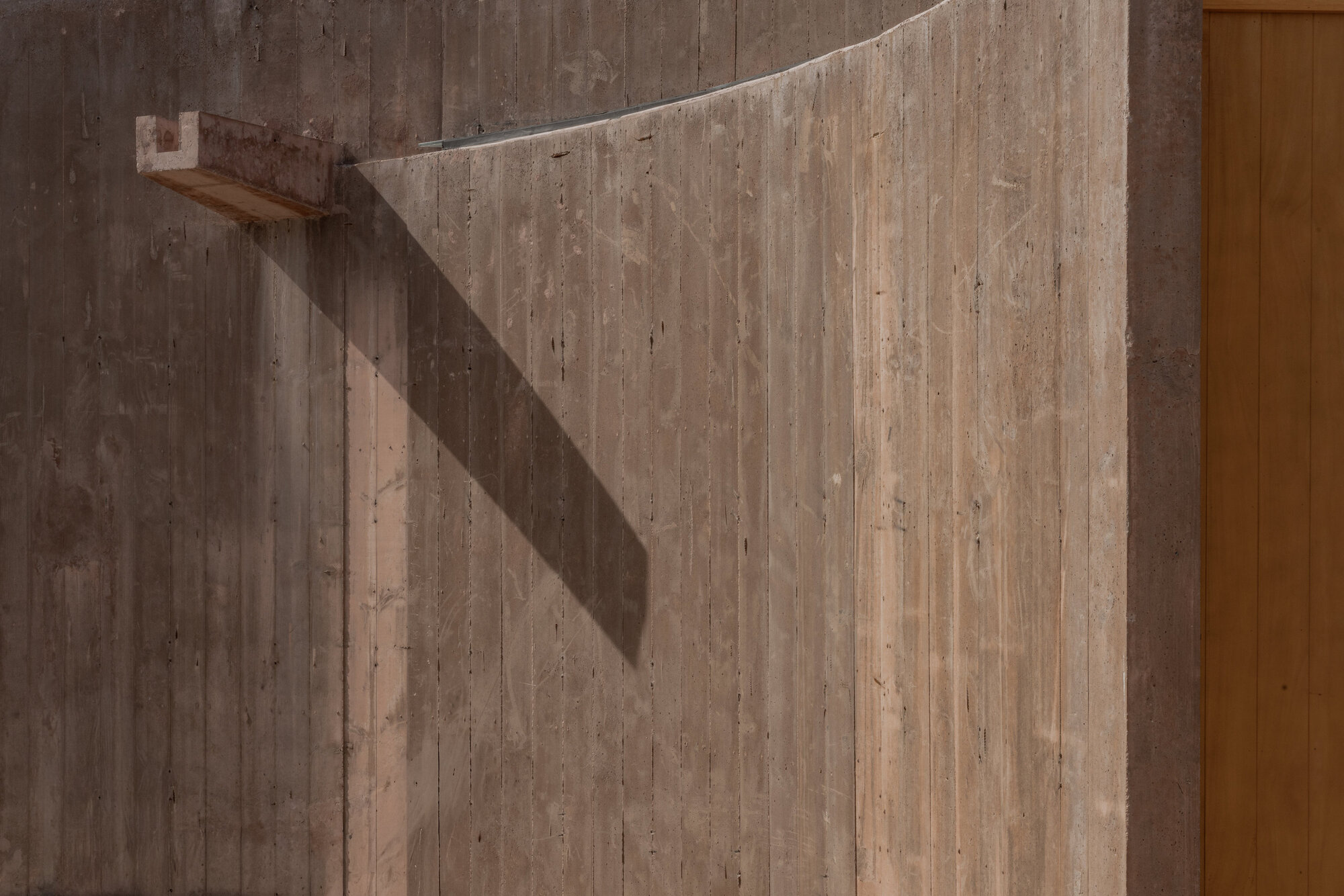 Close-up of vertical board-formed concrete with a projecting light fixture casting a diagonal shadow beside a timber door