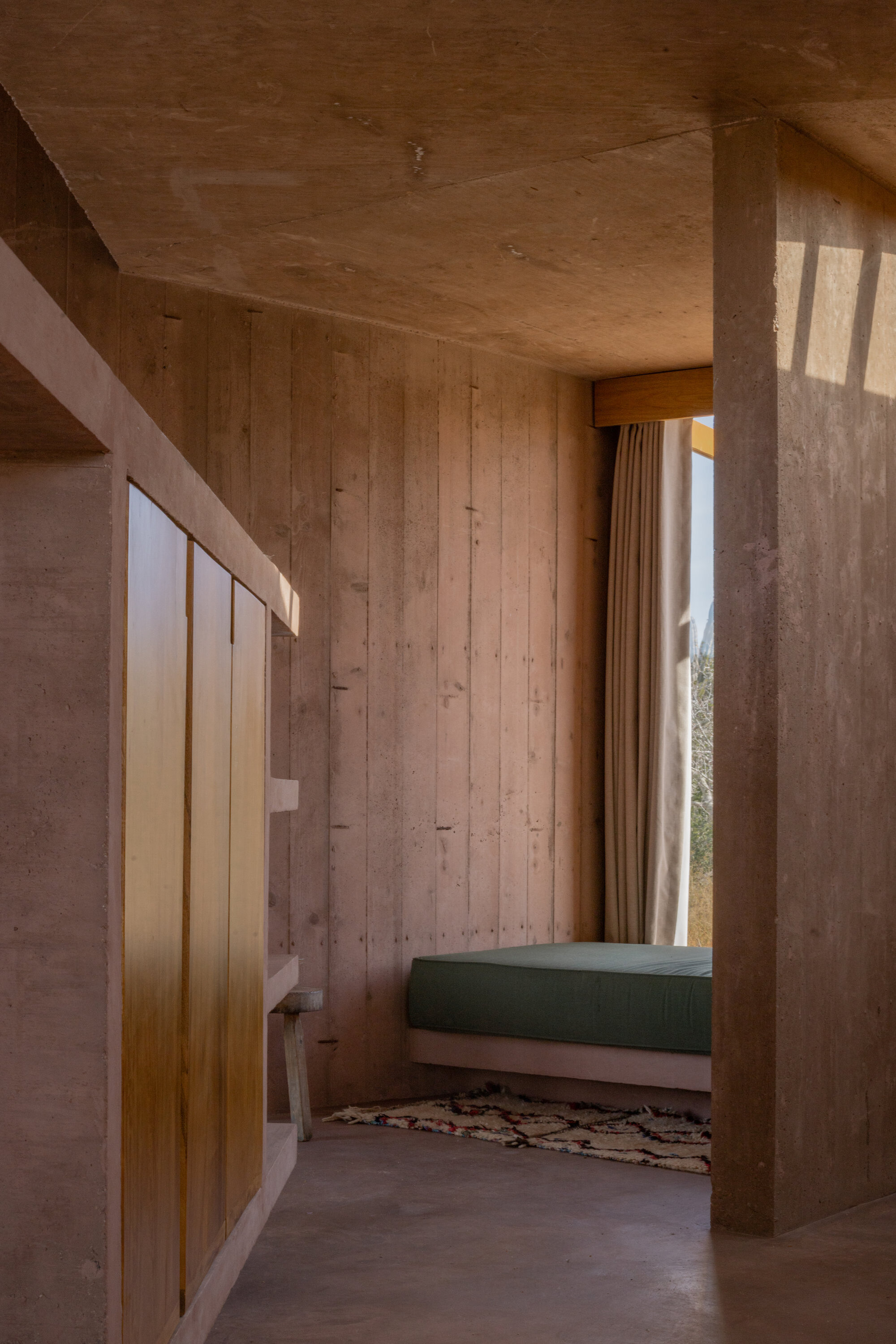 Bedroom nook with board-formed concrete walls, a green daybed and a woven rug, lit by low winter sun through a curtained window
