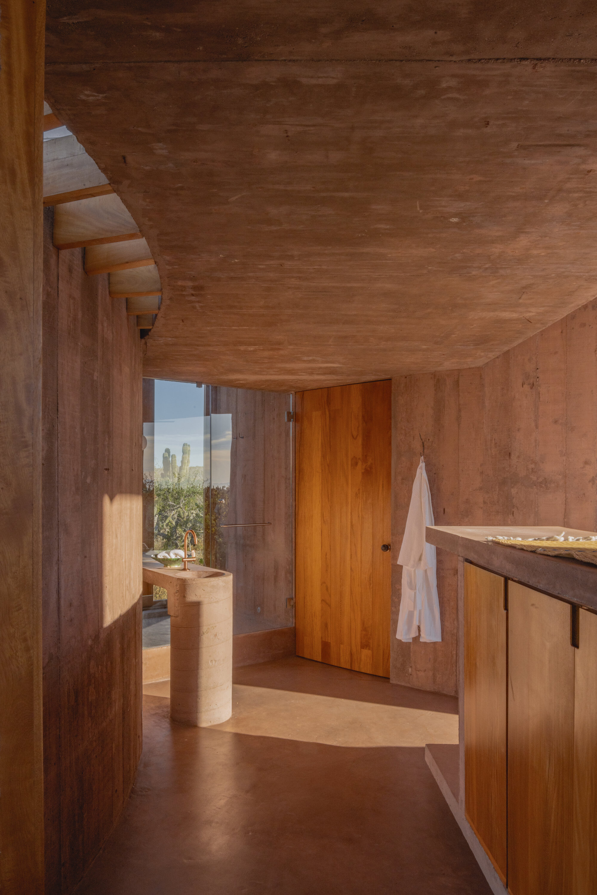 Curved concrete corridor leading to a freestanding stone basin and timber door, with a white robe hanging in warm afternoon light
