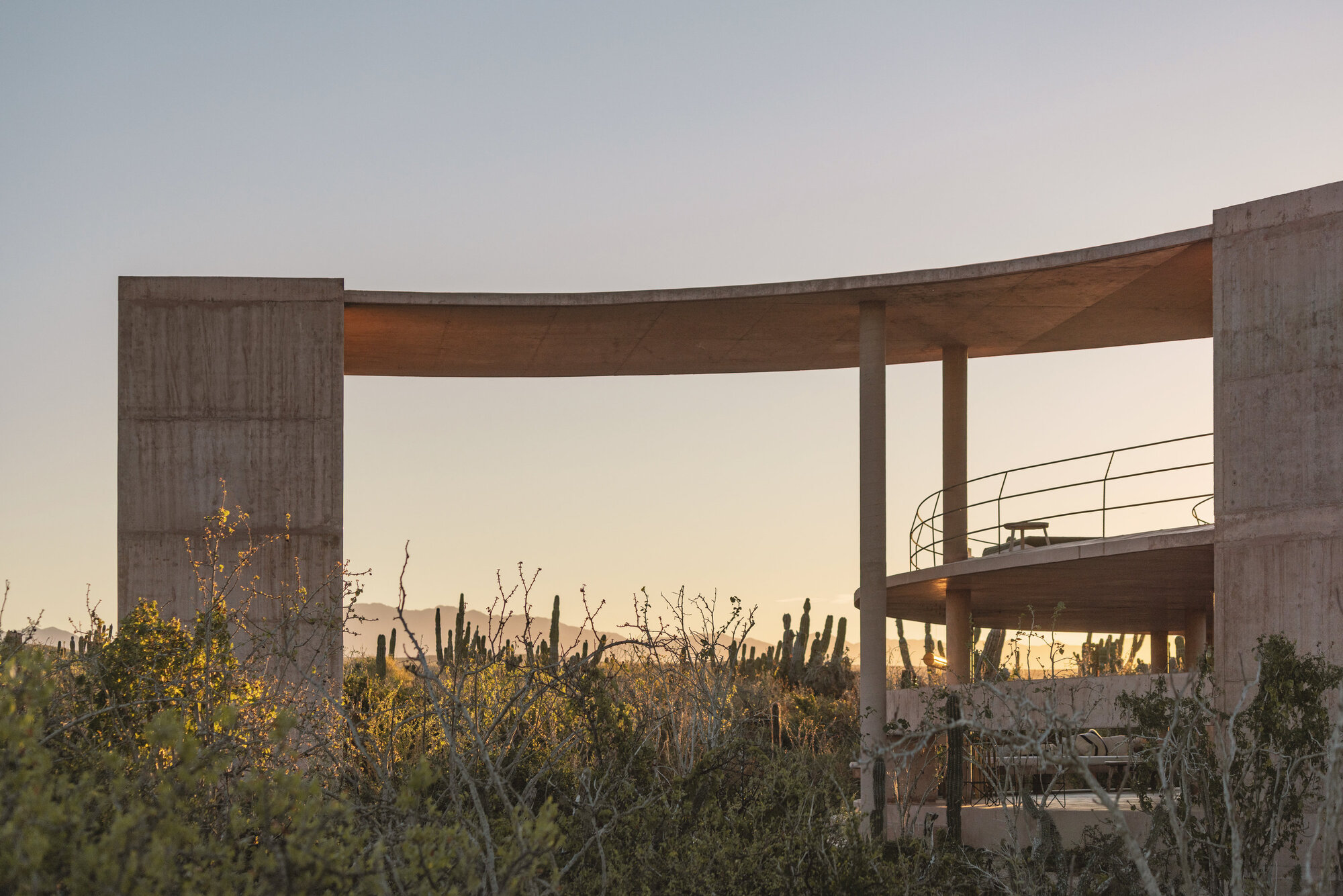 Curved concrete roof slab on thin columns and two massive piers, with a railed upper deck against cactus and a glowing sunset sky