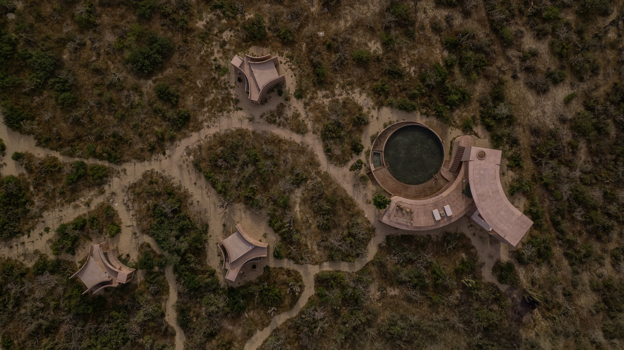 Aerial view of a round pool, curved pavilion roofs and three small cabins linked by sandy paths through dark desert scrub