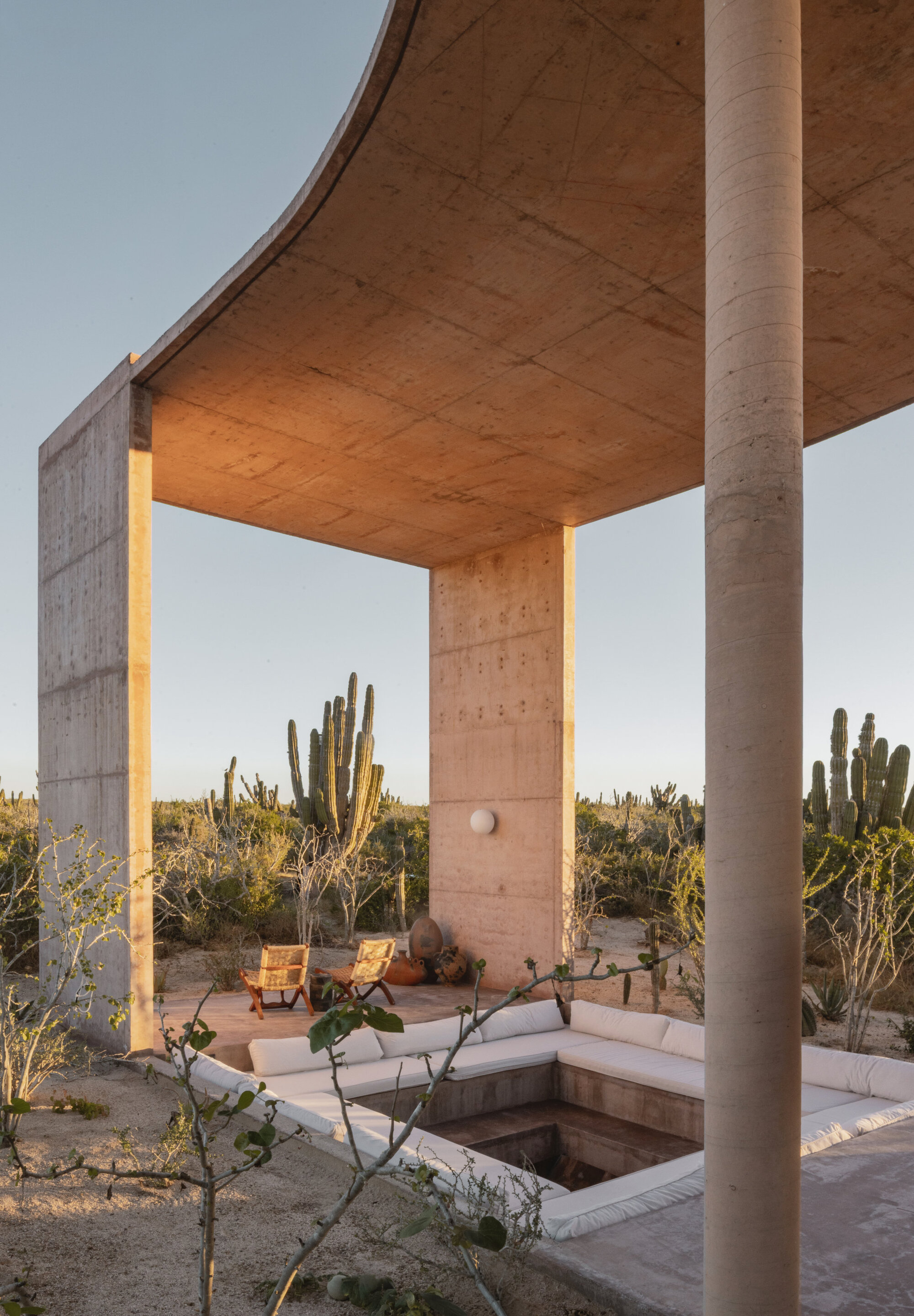 Sunken seating pit with white cushions under a curved concrete canopy, framed by tall cacti and a round column in late light