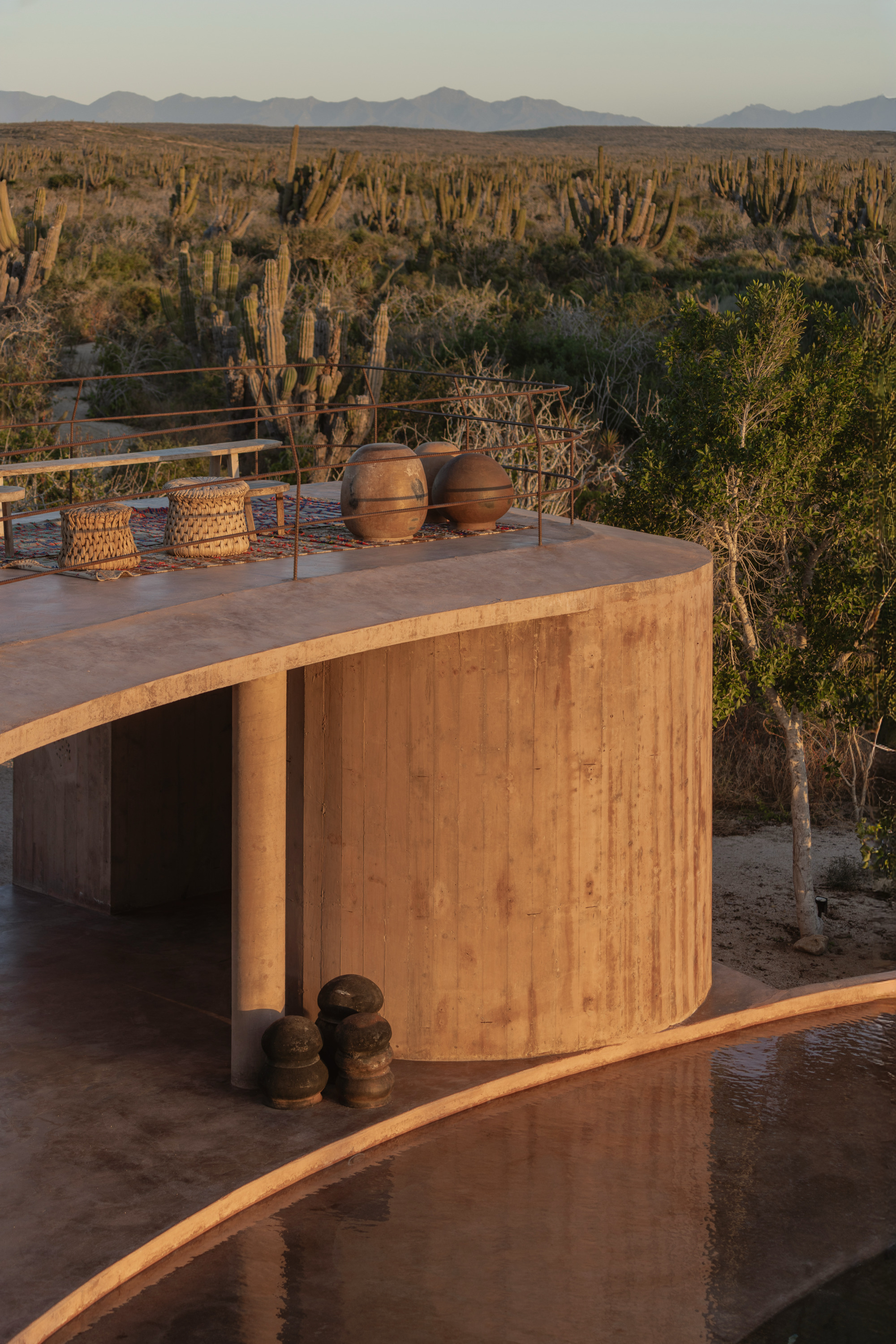 Curved board-formed concrete drum with clay pots on the roof terrace beside a reflecting pool, lit by low desert sun