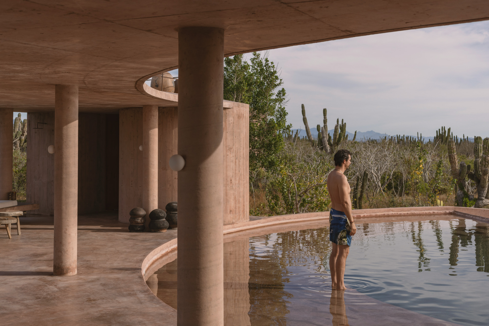 Man standing knee-deep in a curved pool beneath a concrete canopy, with cardon cacti beyond in soft afternoon light