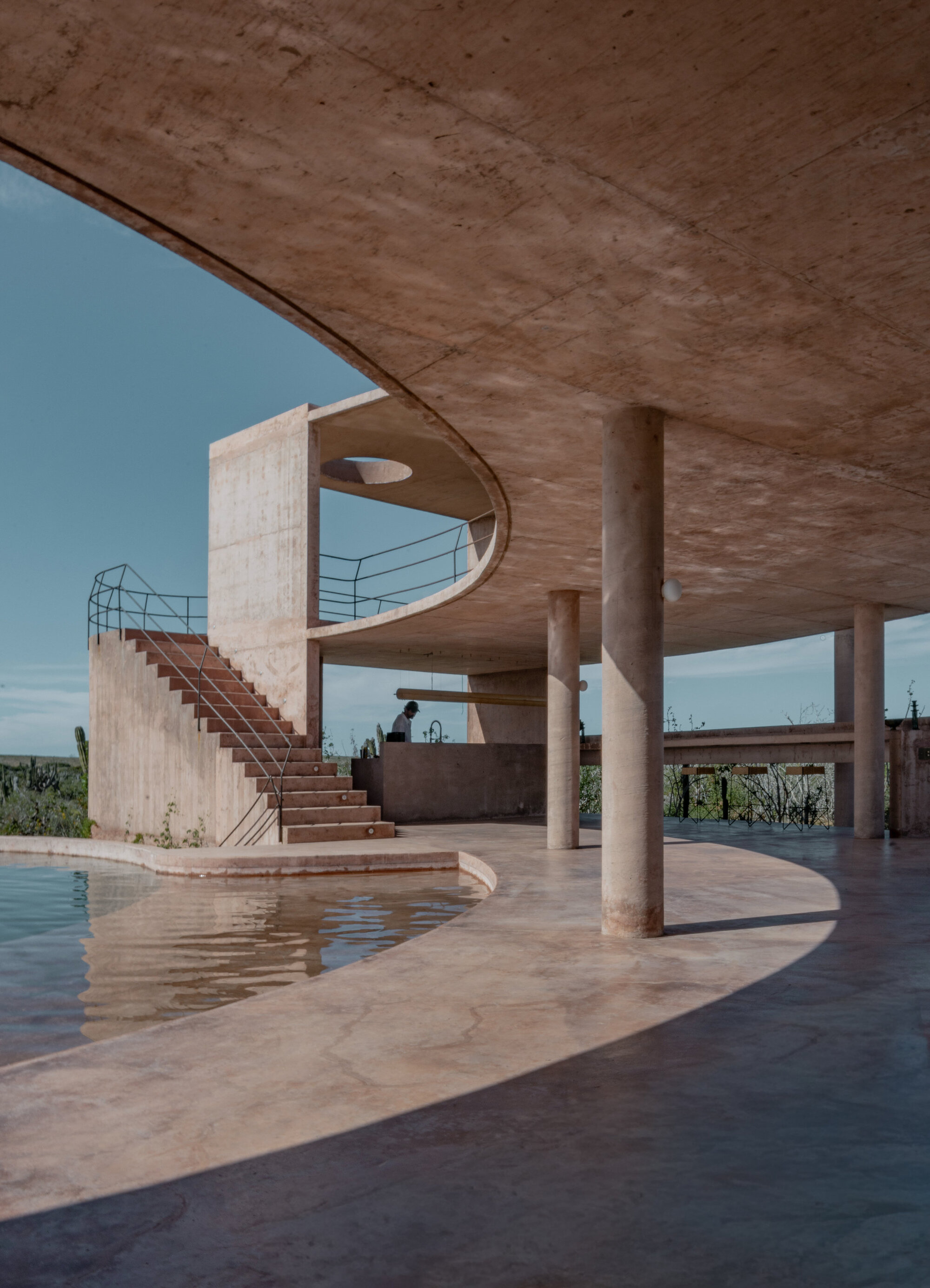 Curving concrete canopy on round columns above a pool edge and exposed stair, under a clear blue sky