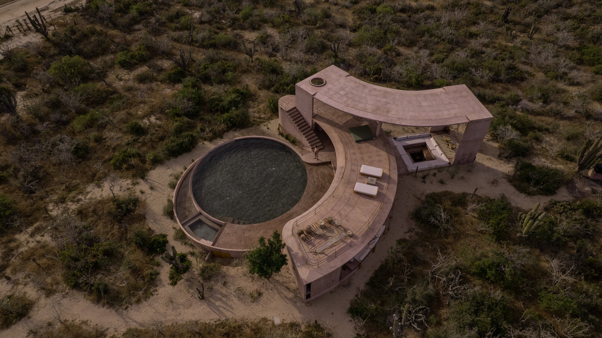 Aerial view of the crescent-shaped rammed earth structure beside a round pool, ringed by desert scrub in dim evening light
