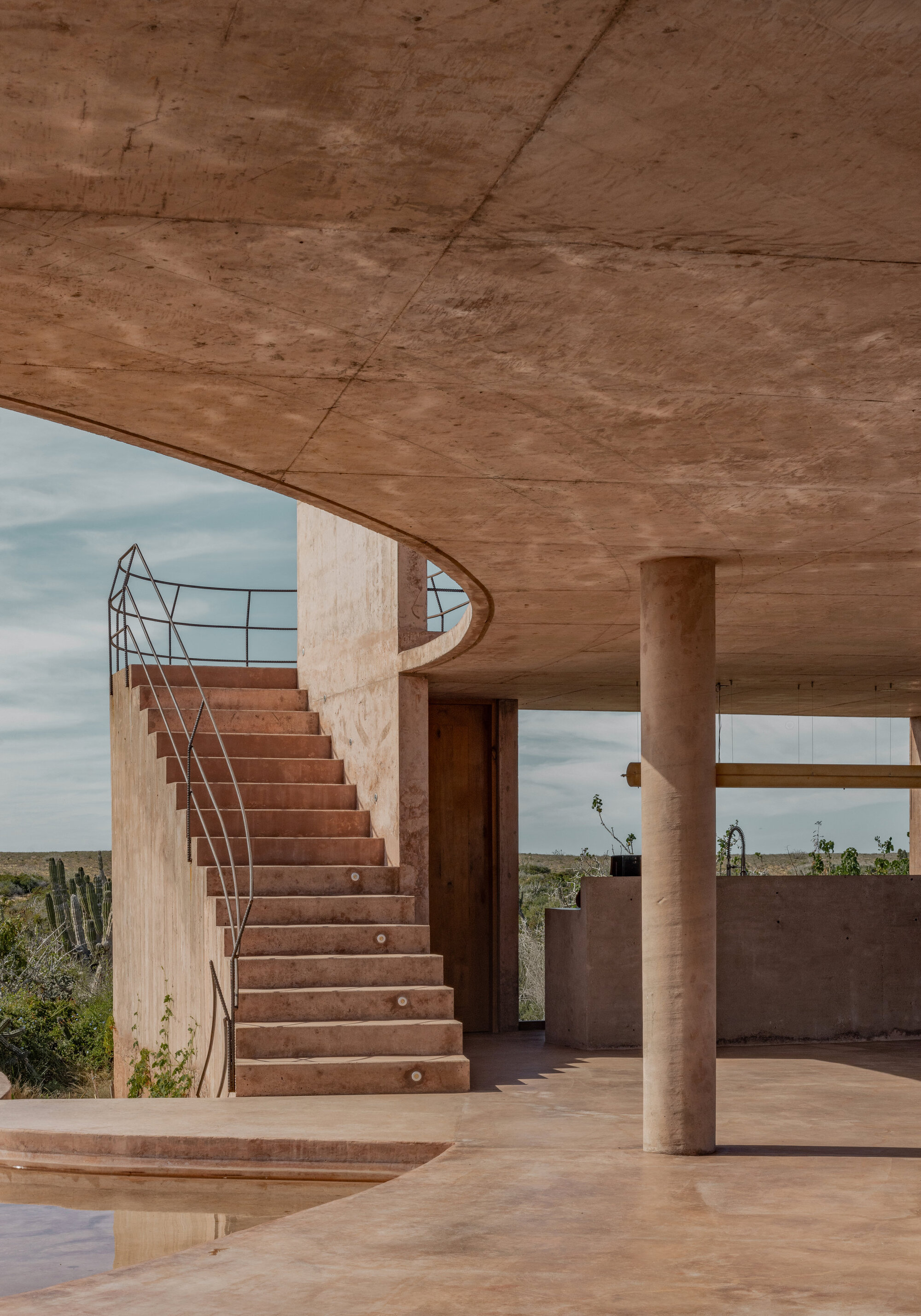 Rammed earth stair with metal railing rising beside a round column under a curved concrete canopy, cactus visible beyond