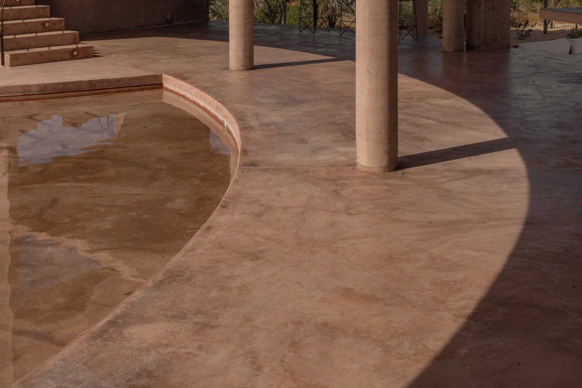 Curved pool edge and pink concrete floor with round columns casting a sharp arc of shadow in low sun