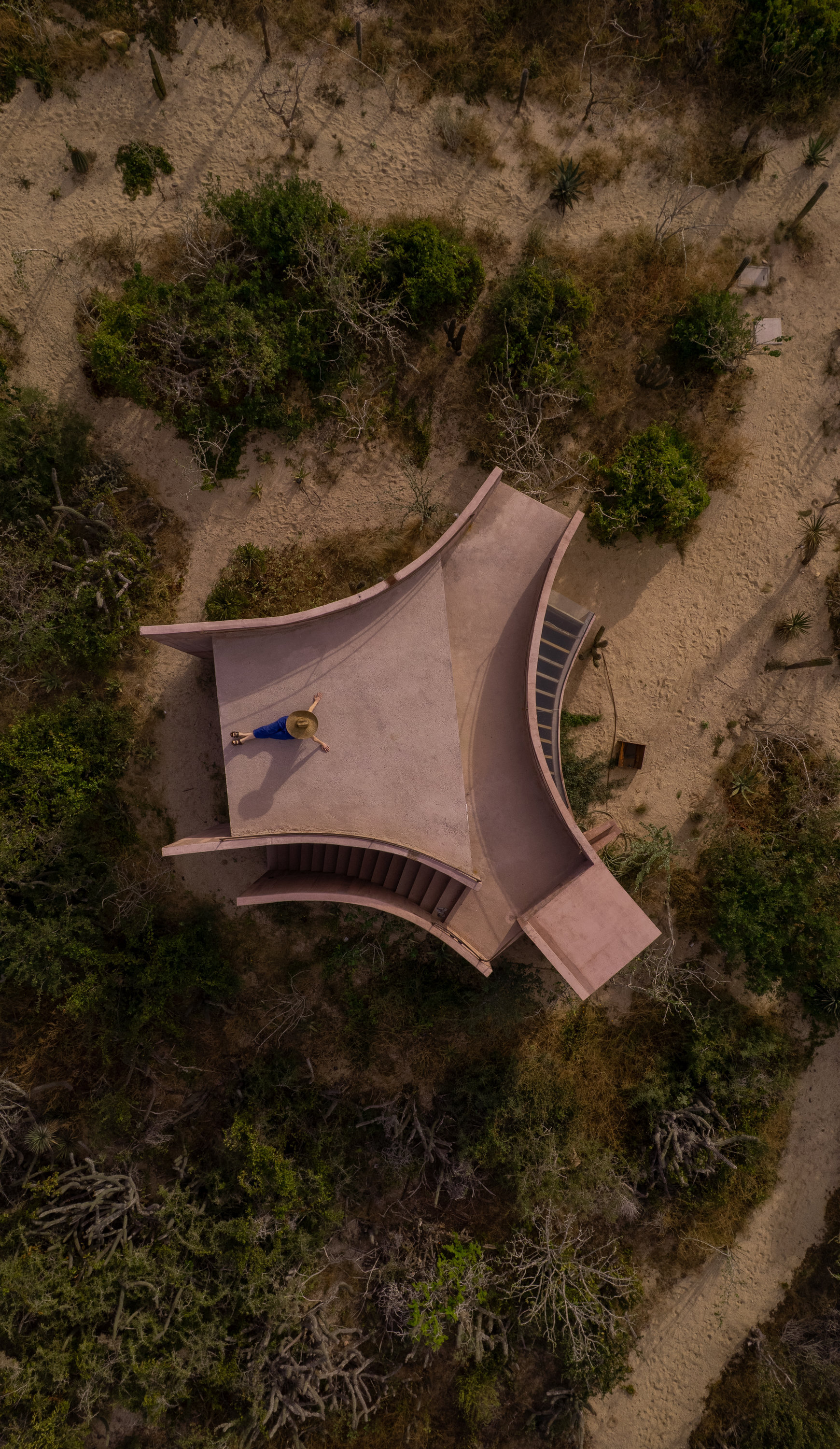 Top-down view of the star-shaped pink roof with a person lying on it, surrounded by sandy paths and shrubs