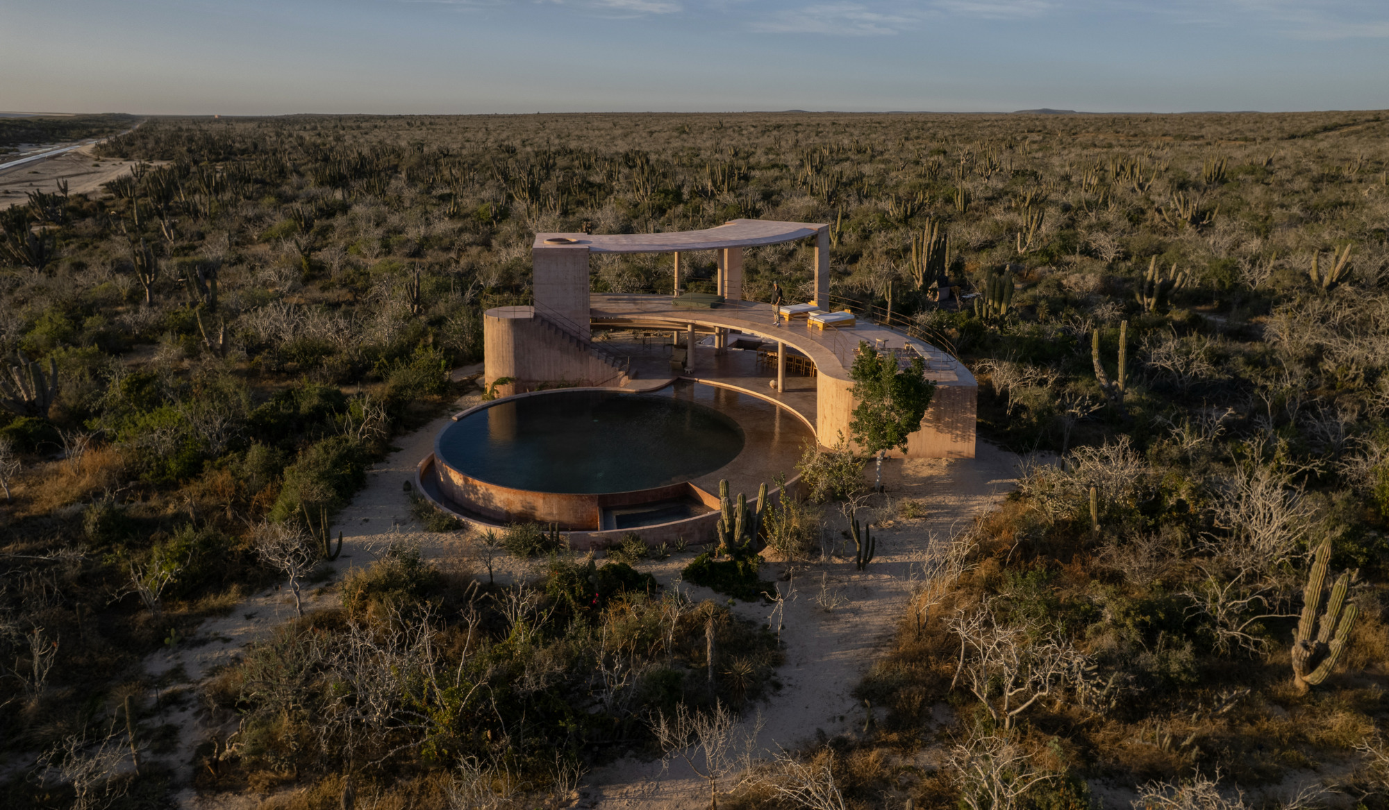 Low aerial view of the curved pavilion and circular pool in a flat cactus desert at sunset