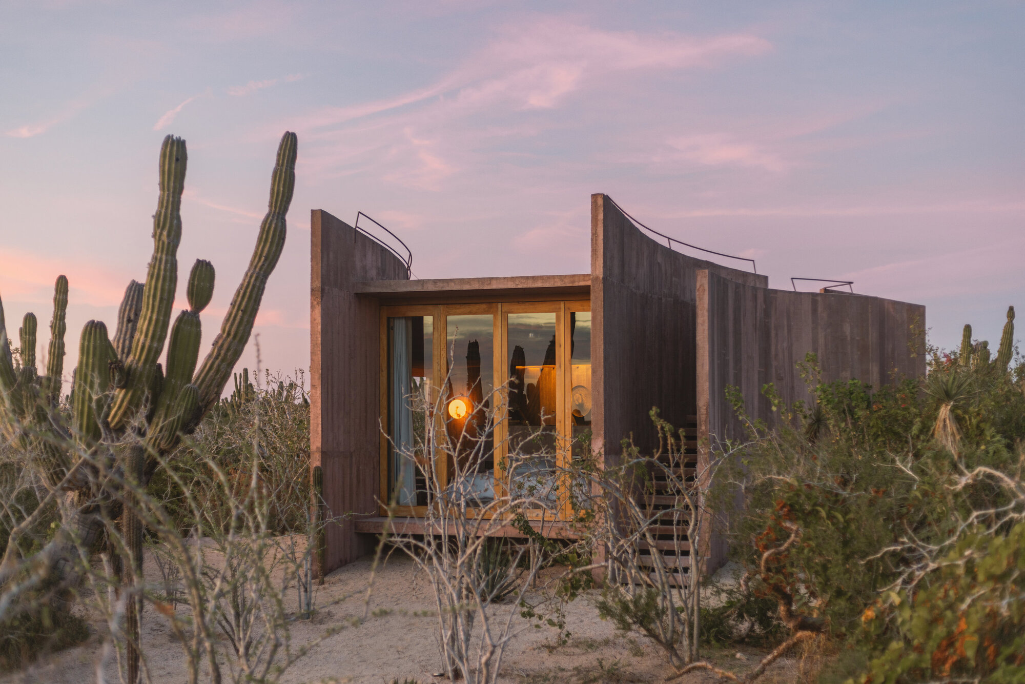 Glazed cabin front with a glowing wall lamp between curved board-formed walls, seen among cacti at pink dusk