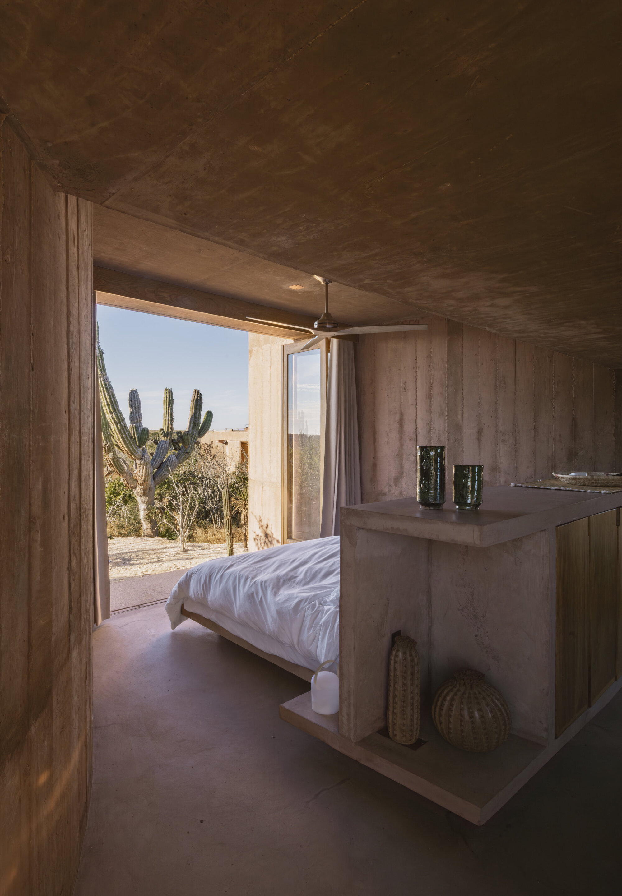 Bedroom interior with board-formed walls and ceiling, a white bed and shelf unit, opening onto a cactus in daylight