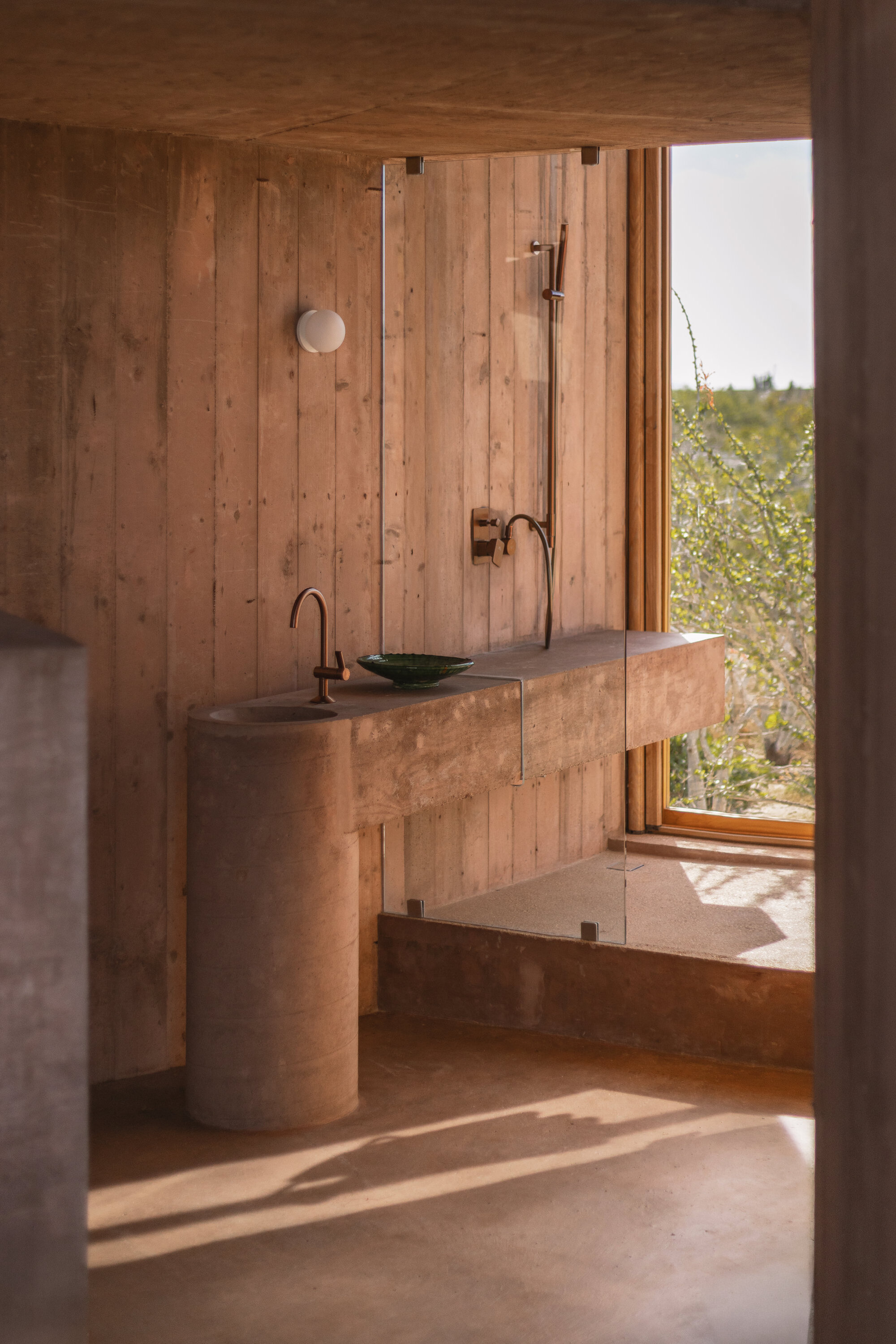 Bathroom with a cylindrical stone pedestal sink, green basin and glass-screened shower, streaked by afternoon sunlight on the floor
