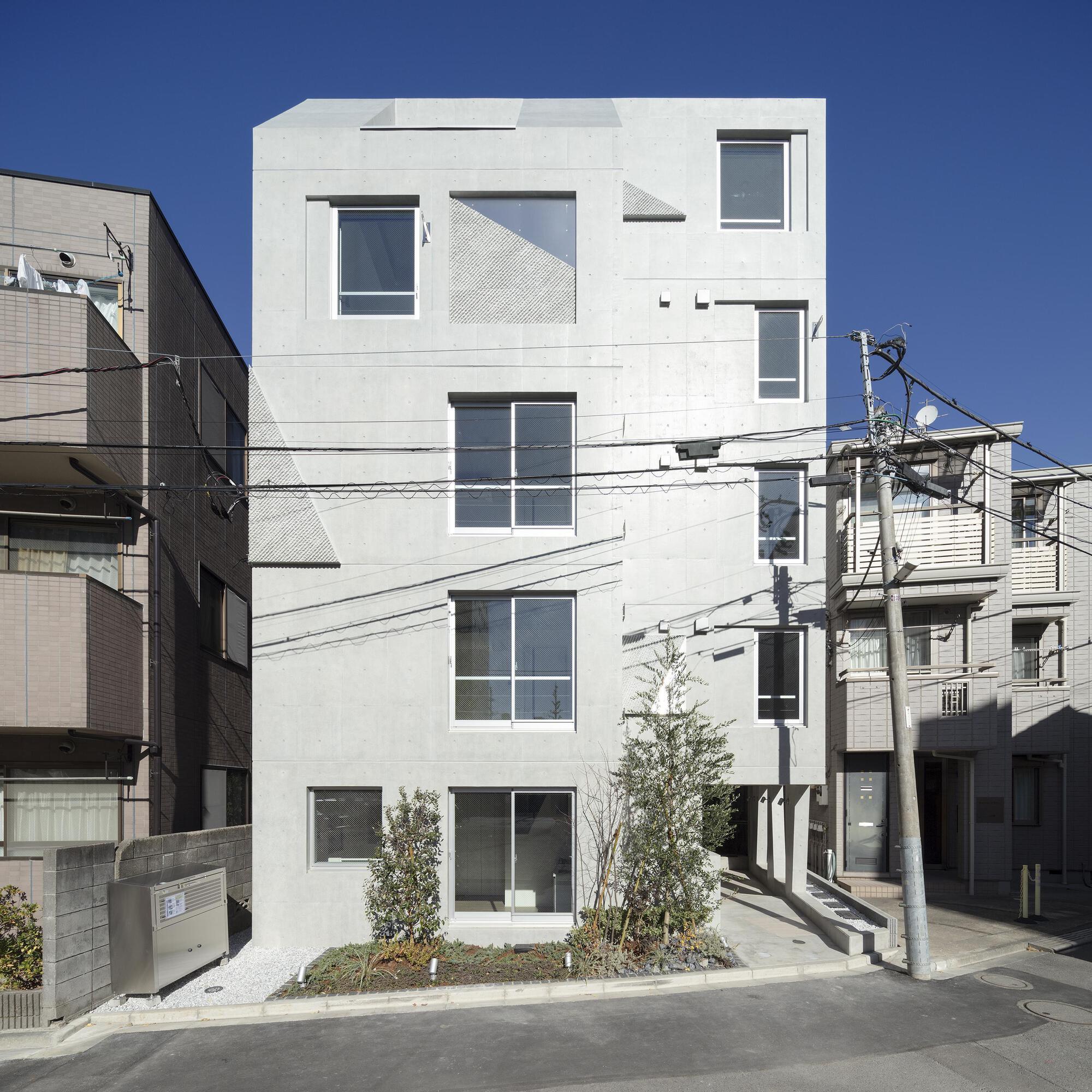 Front facade of the board-formed concrete apartment block with irregular windows and small planted bed, under a clear blue winter sky