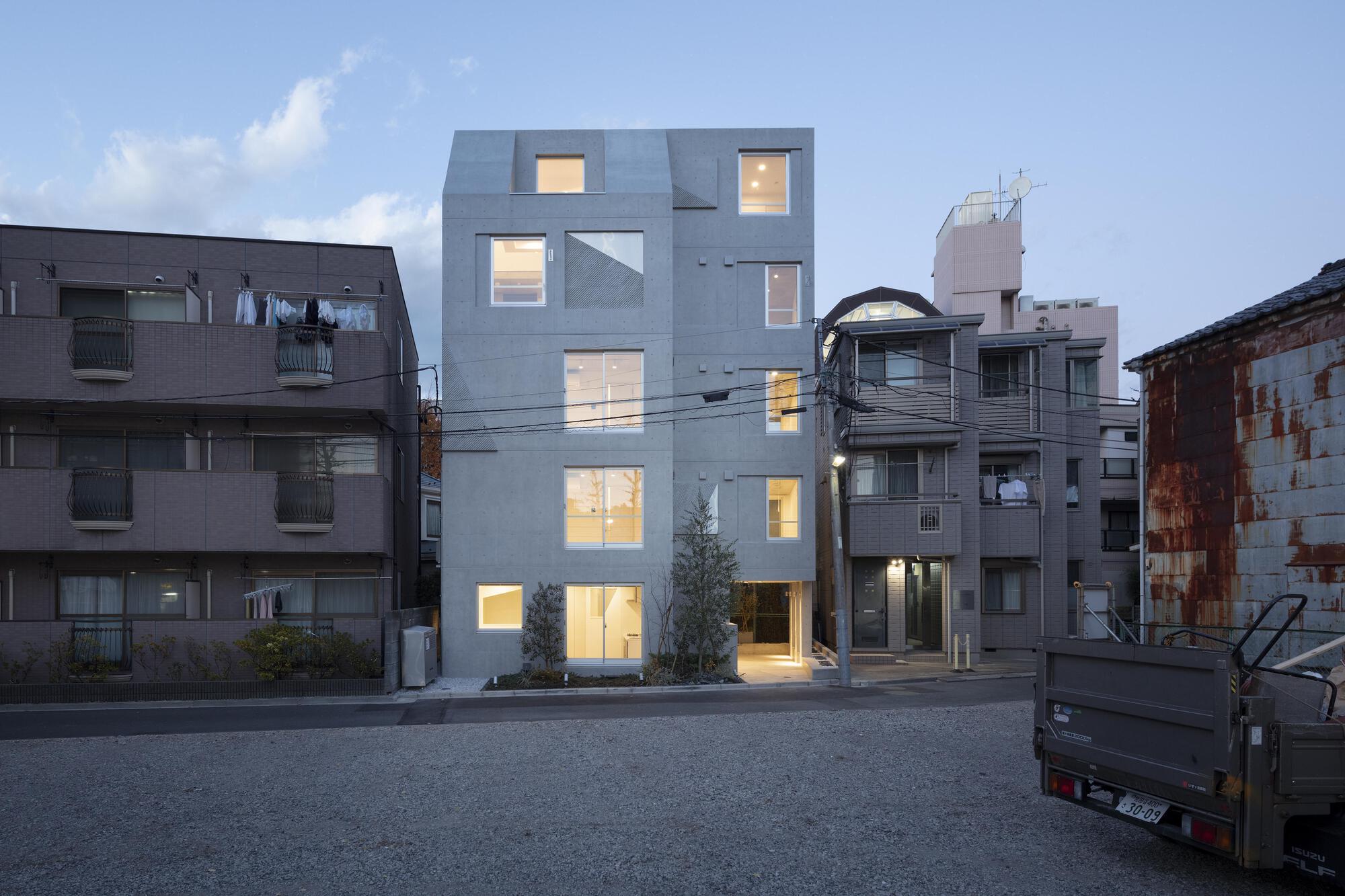 Concrete apartment block at dusk with warm light glowing in its windows, framed by neighbouring houses and a parked truck