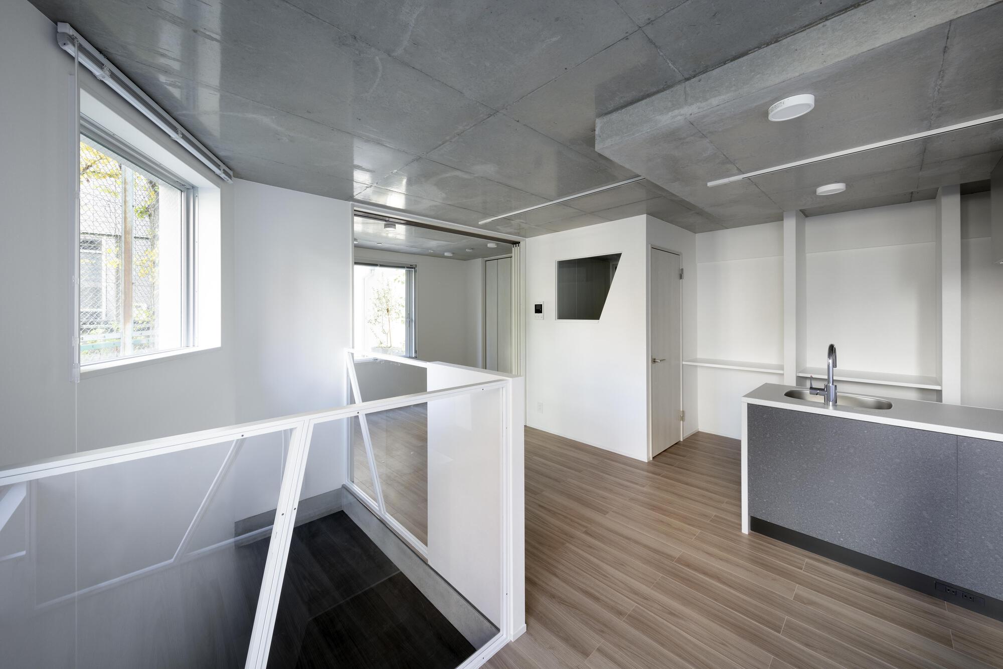 Open living level with exposed concrete ceiling, glass stair balustrade and a grey kitchen counter, lit by a side window