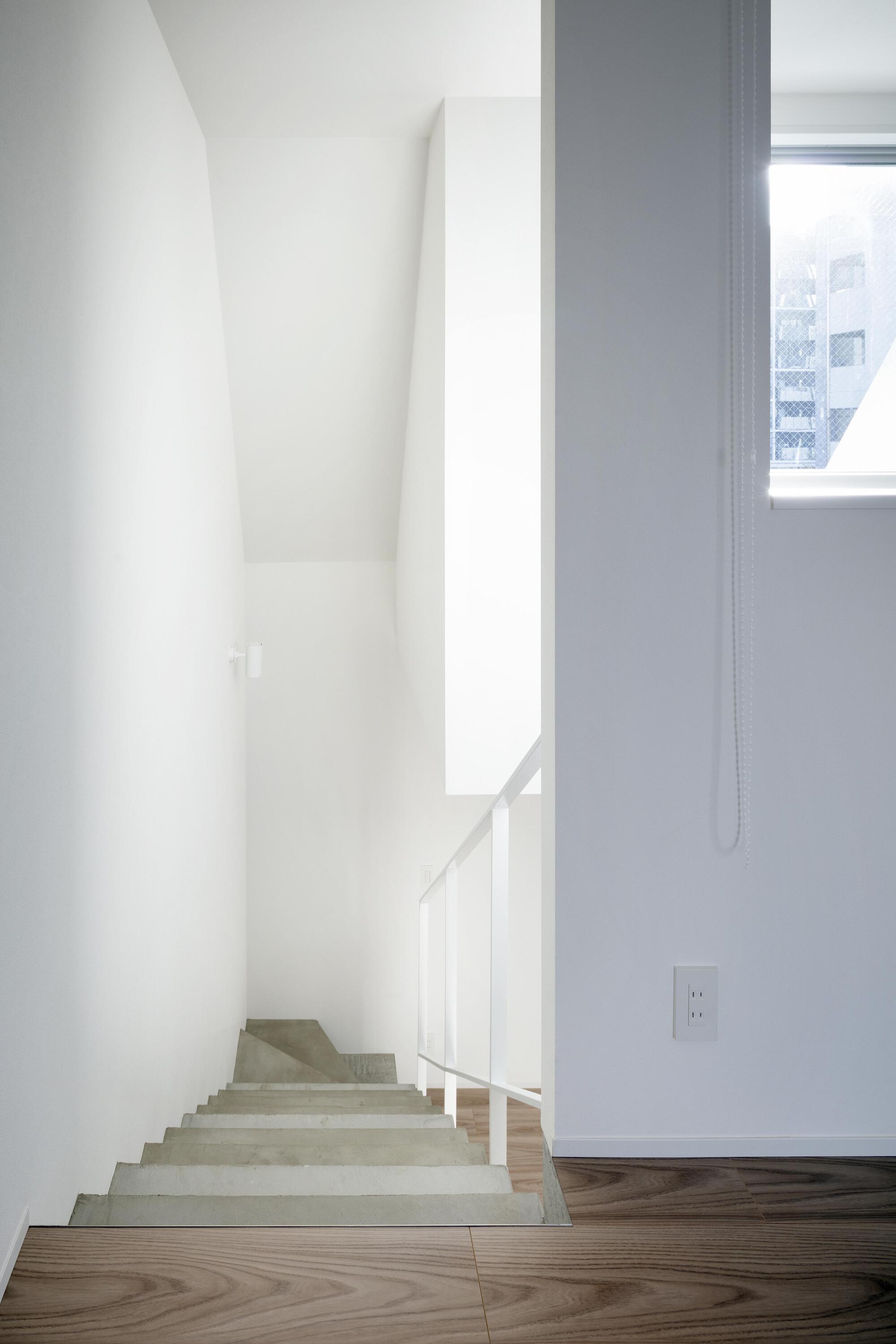 Narrow white stairwell with concrete treads descending from a timber floor, a window with daylight at right