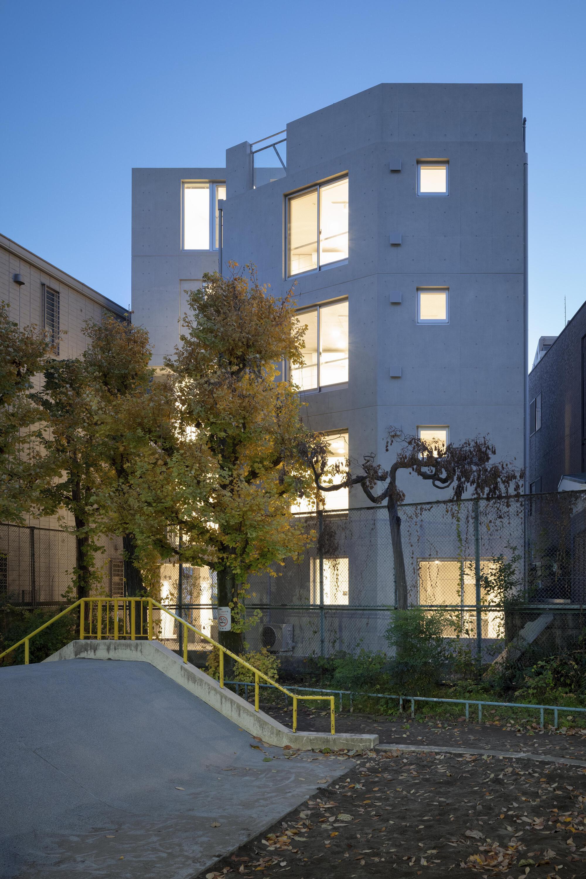 Board-formed concrete apartment block with lit windows at dusk, seen over a yellow-railed ramp and autumn trees