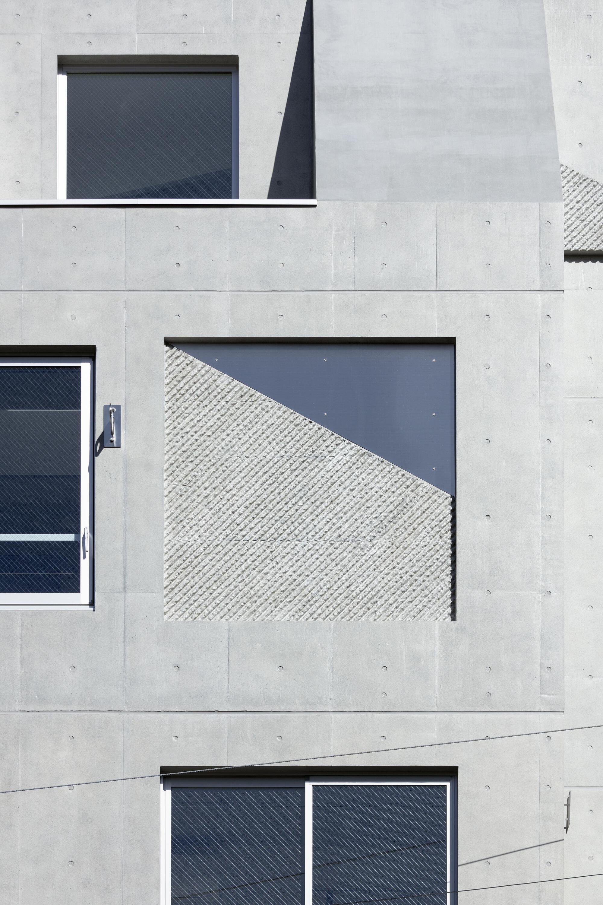 Facade detail of a square window opening filled half with textured gray panel, set in concrete with form-tie holes