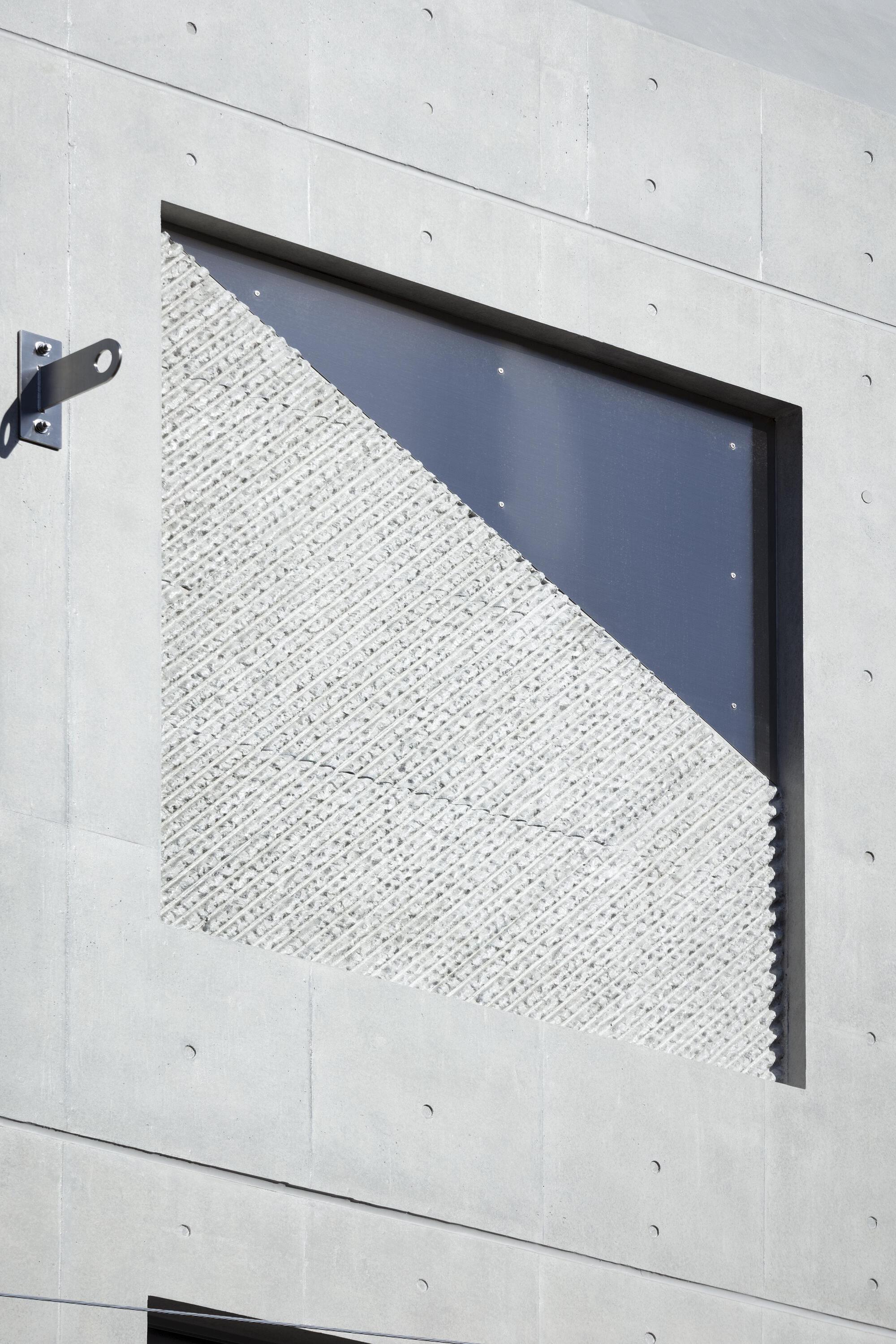 Close-up of a square window recess with a diagonal split between ribbed stone-like panel and dark glass in sunlight
