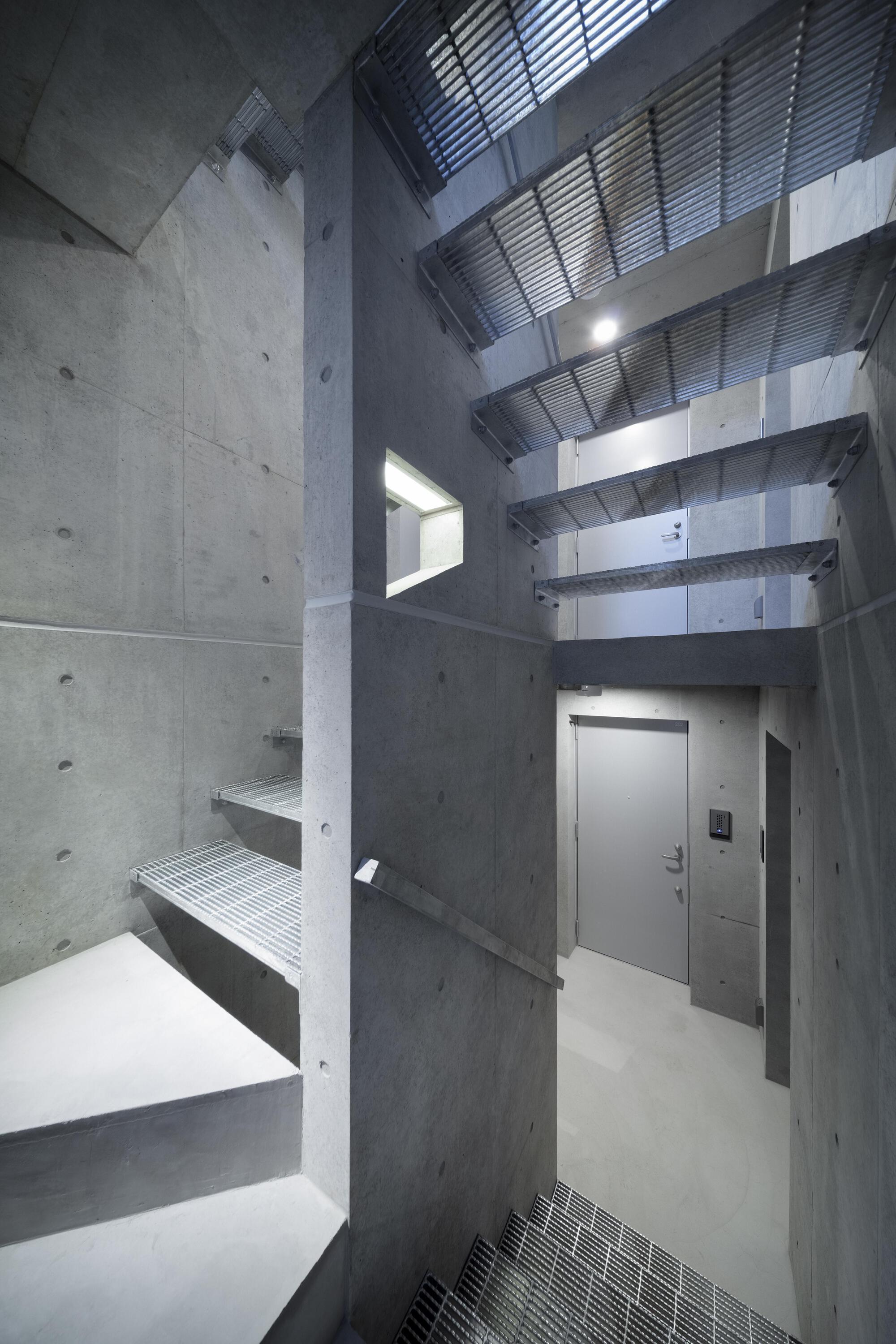 Concrete stairwell with open steel grating treads climbing toward a skylight, a gray door at the landing