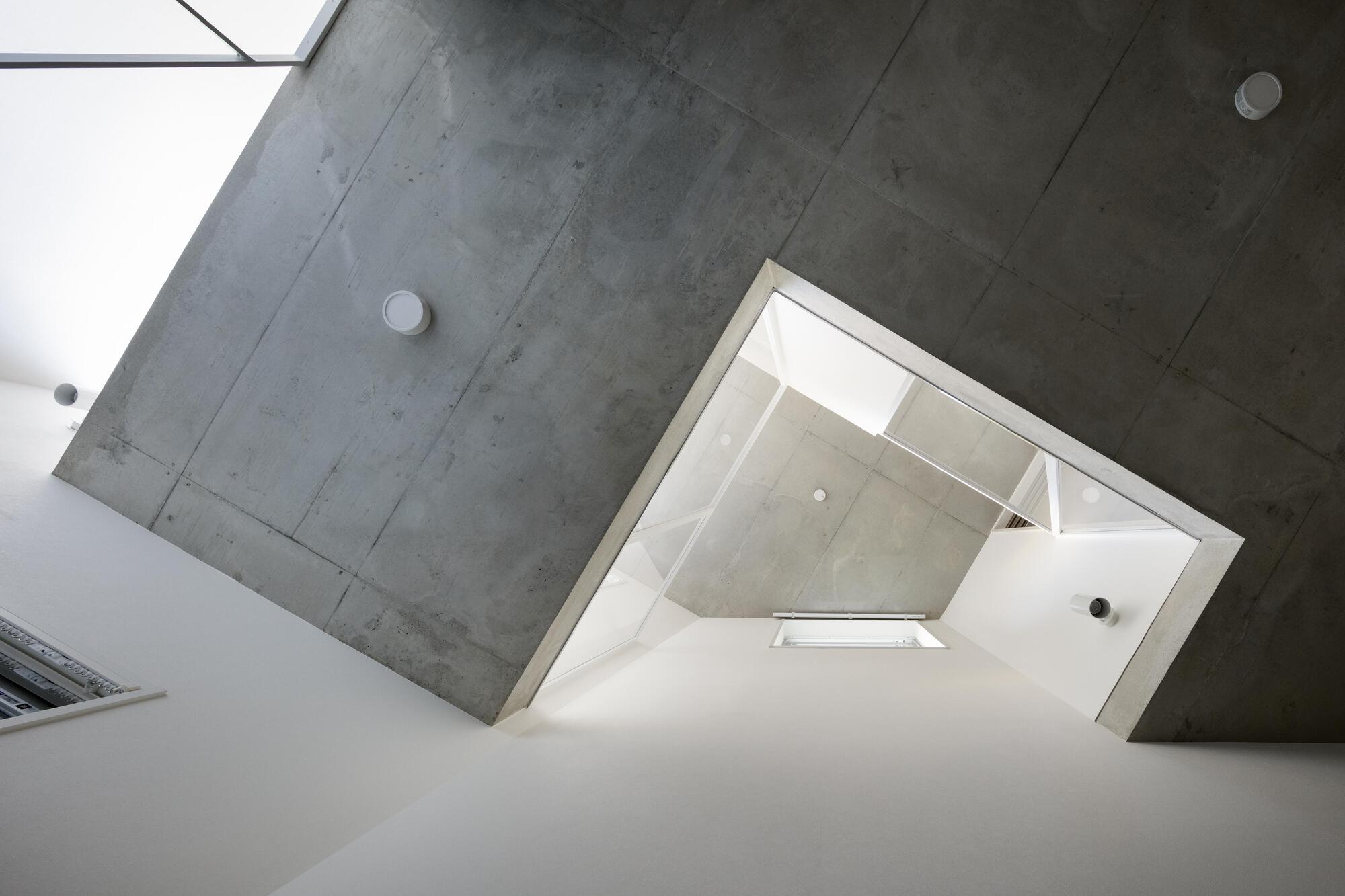 Upward view of a concrete ceiling and angled shaft opening to white walls and a skylight, with round fixtures
