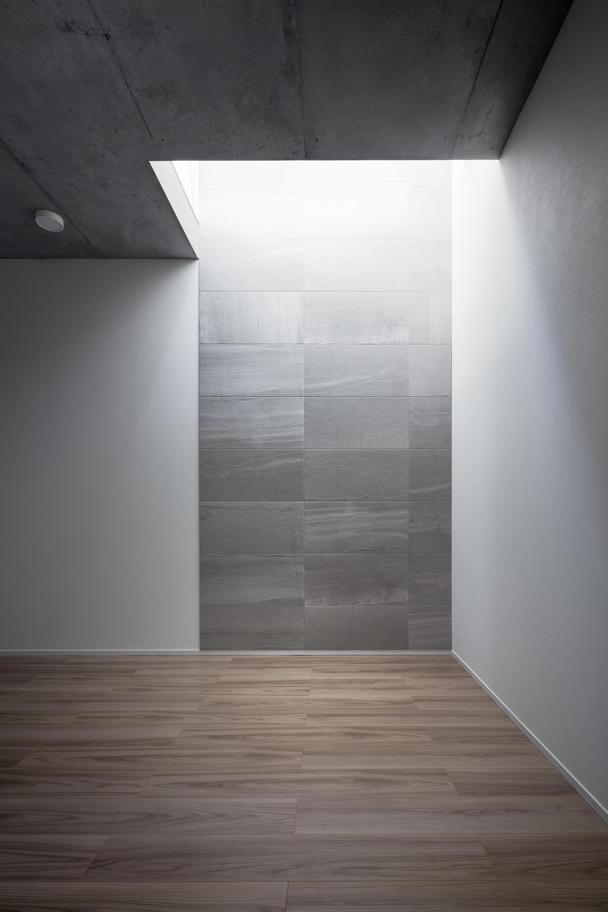 Empty room with wood plank floor, white walls and a tiled gray stone wall lit by a skylight slot above