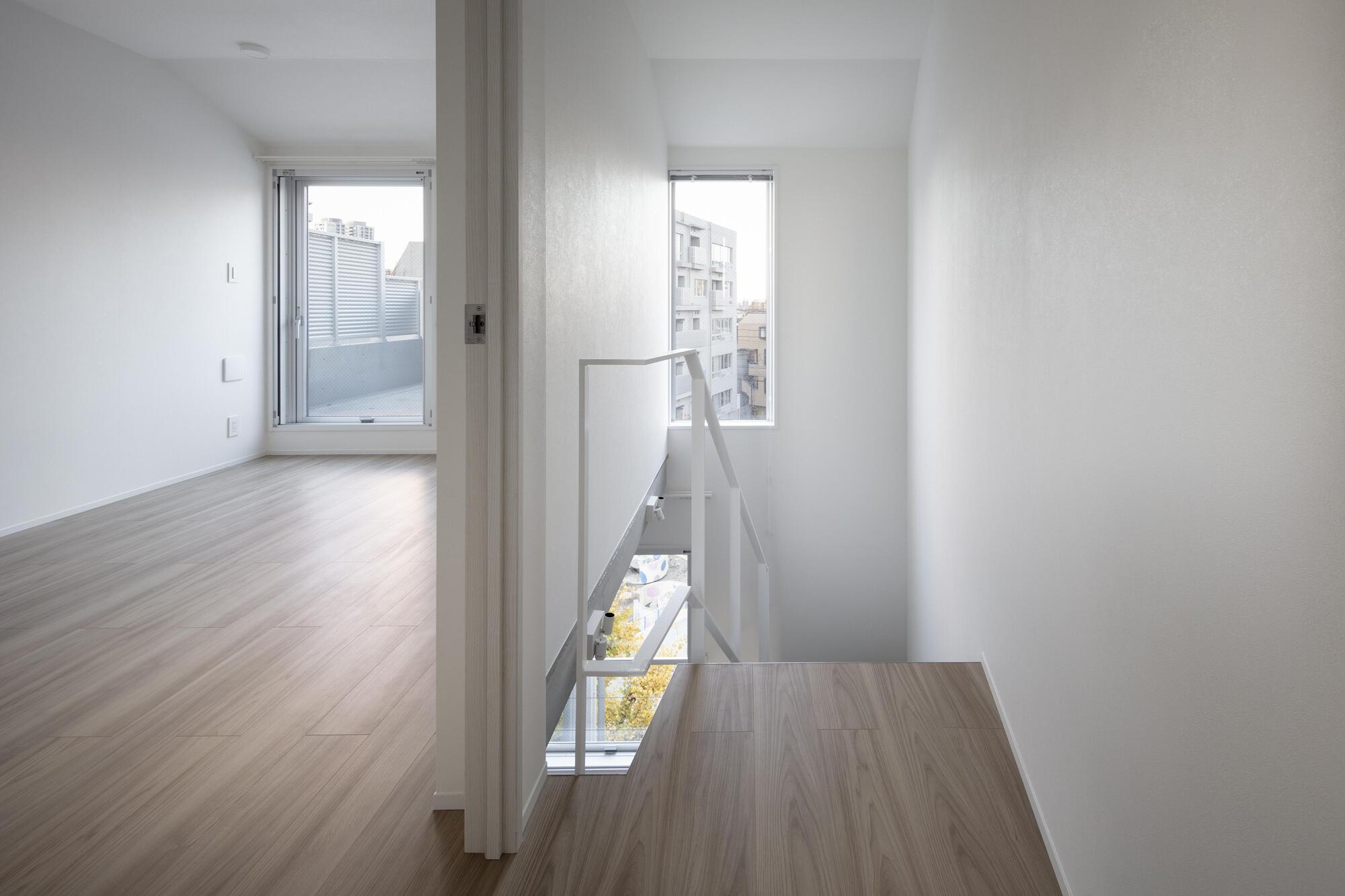 Upper landing with a white railing at the stairwell opening, beside a room whose window looks onto neighboring apartment blocks in daylight