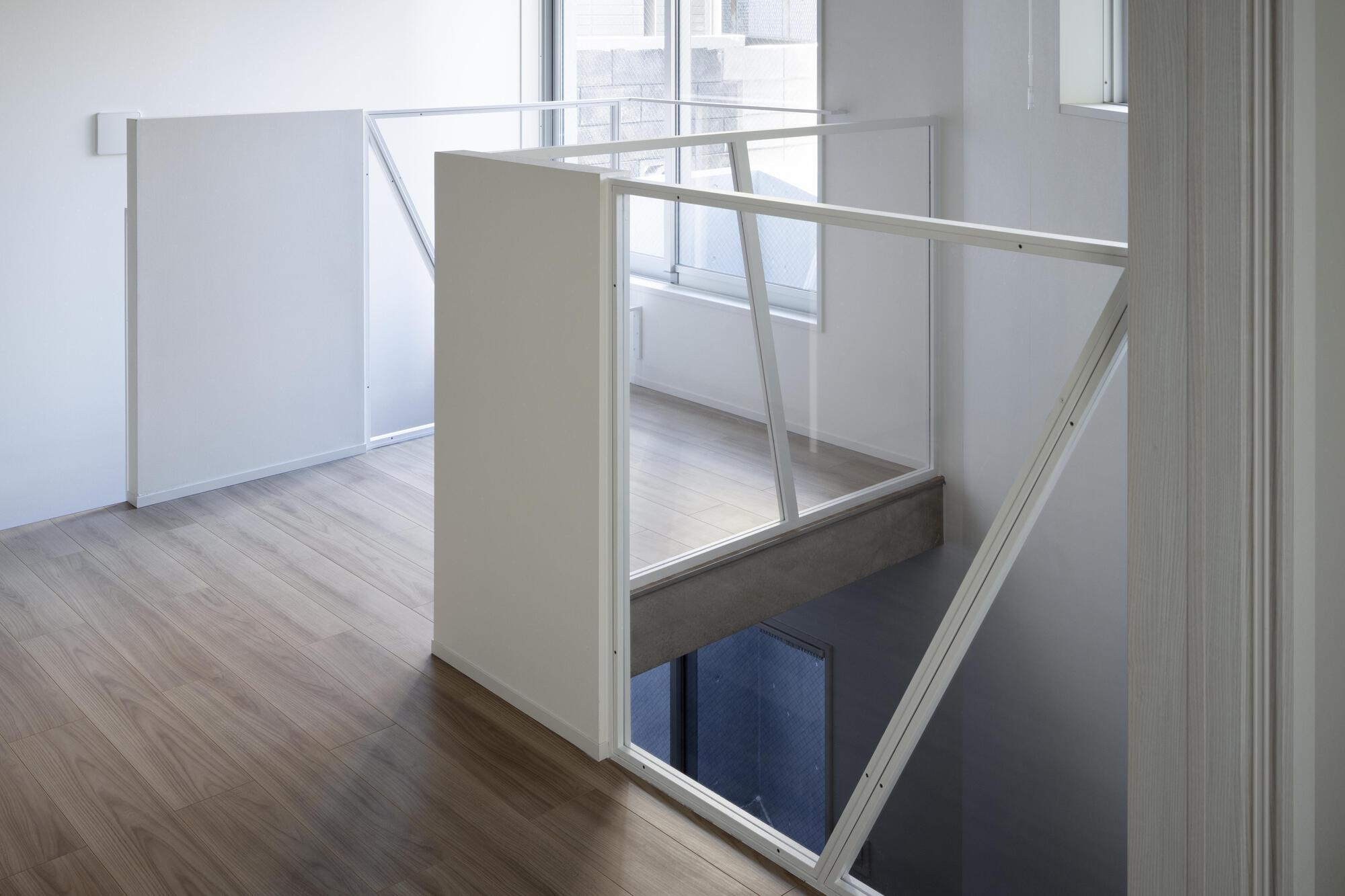 Stair opening edged by angled white metal and glass guards, with a concrete beam below and daylight at the windows
