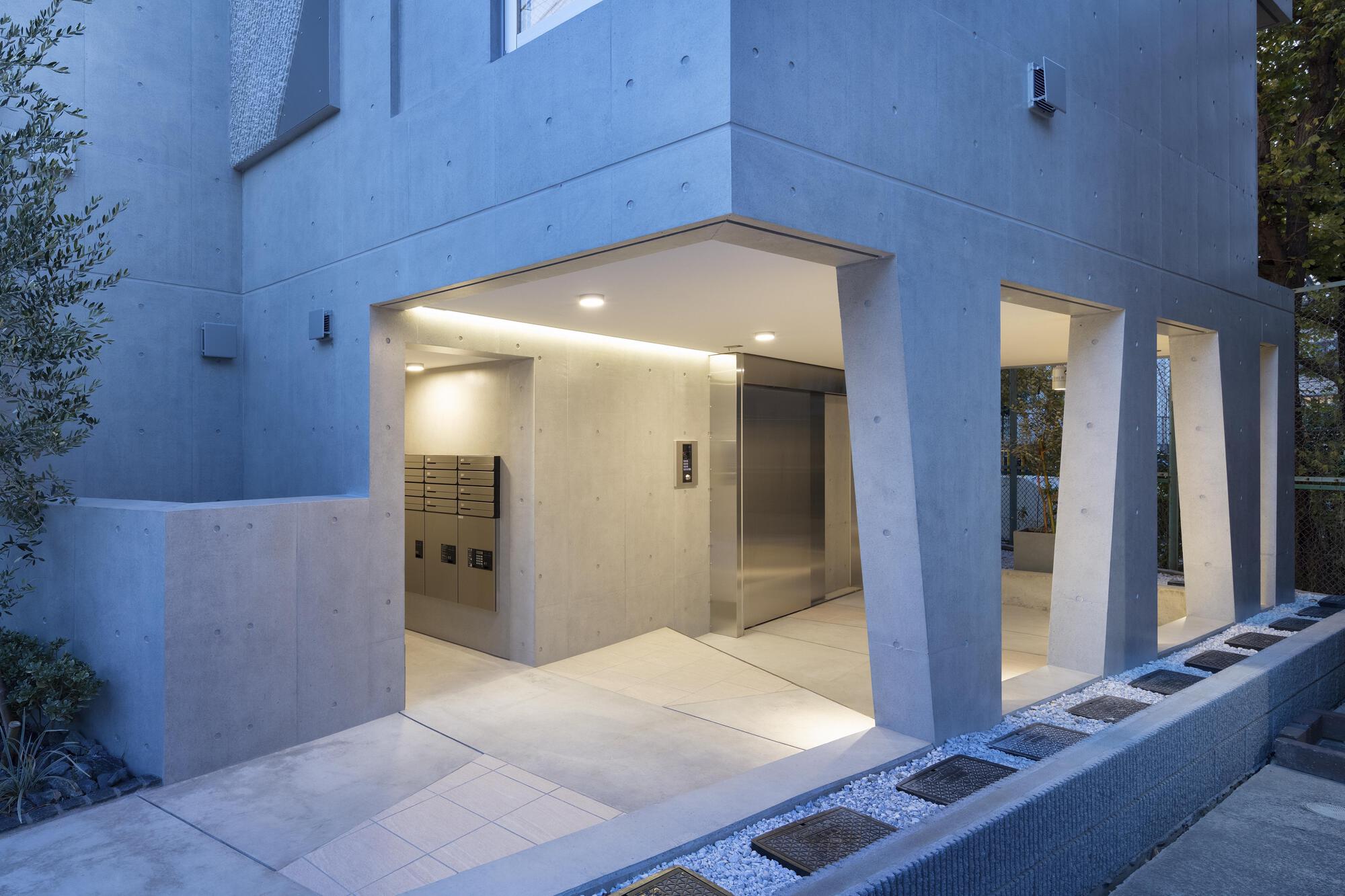 Concrete entrance with slanted piers, mailboxes and a steel elevator door, lit warmly at dusk beside a gravel strip with stepping plates