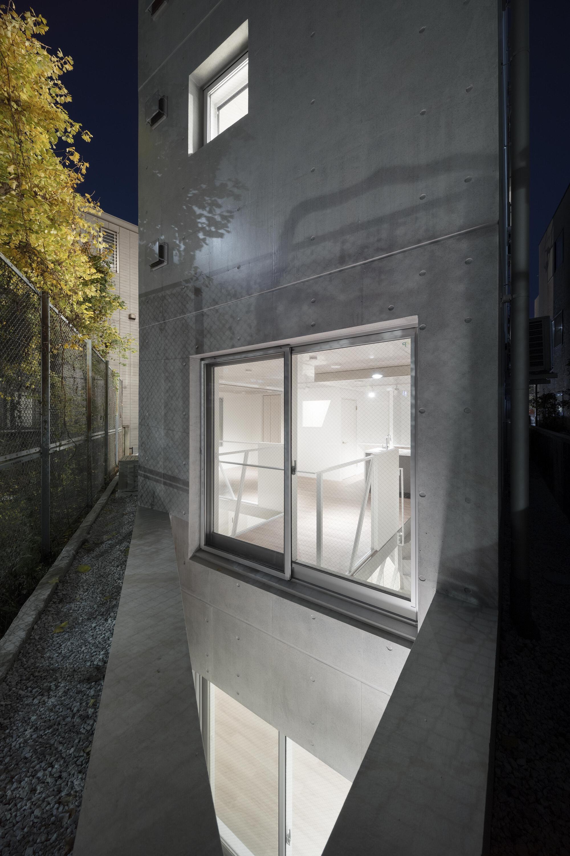 Night view of a narrow concrete facade with a lit sliding window, a basement window below and yellow leaves beside a fence