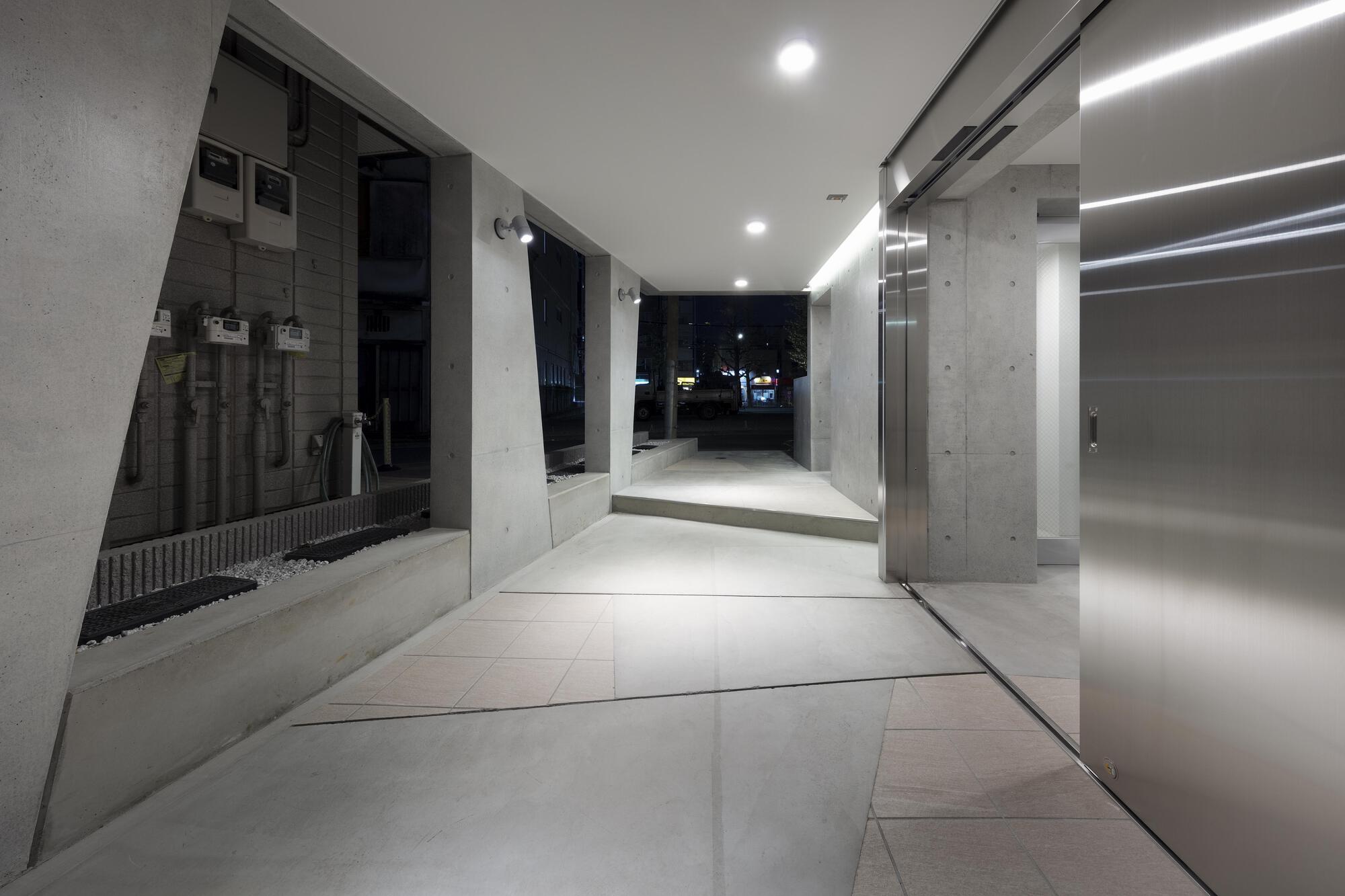 Entrance passage at night with angled concrete piers, utility meters on the left and a brushed steel elevator door on the right