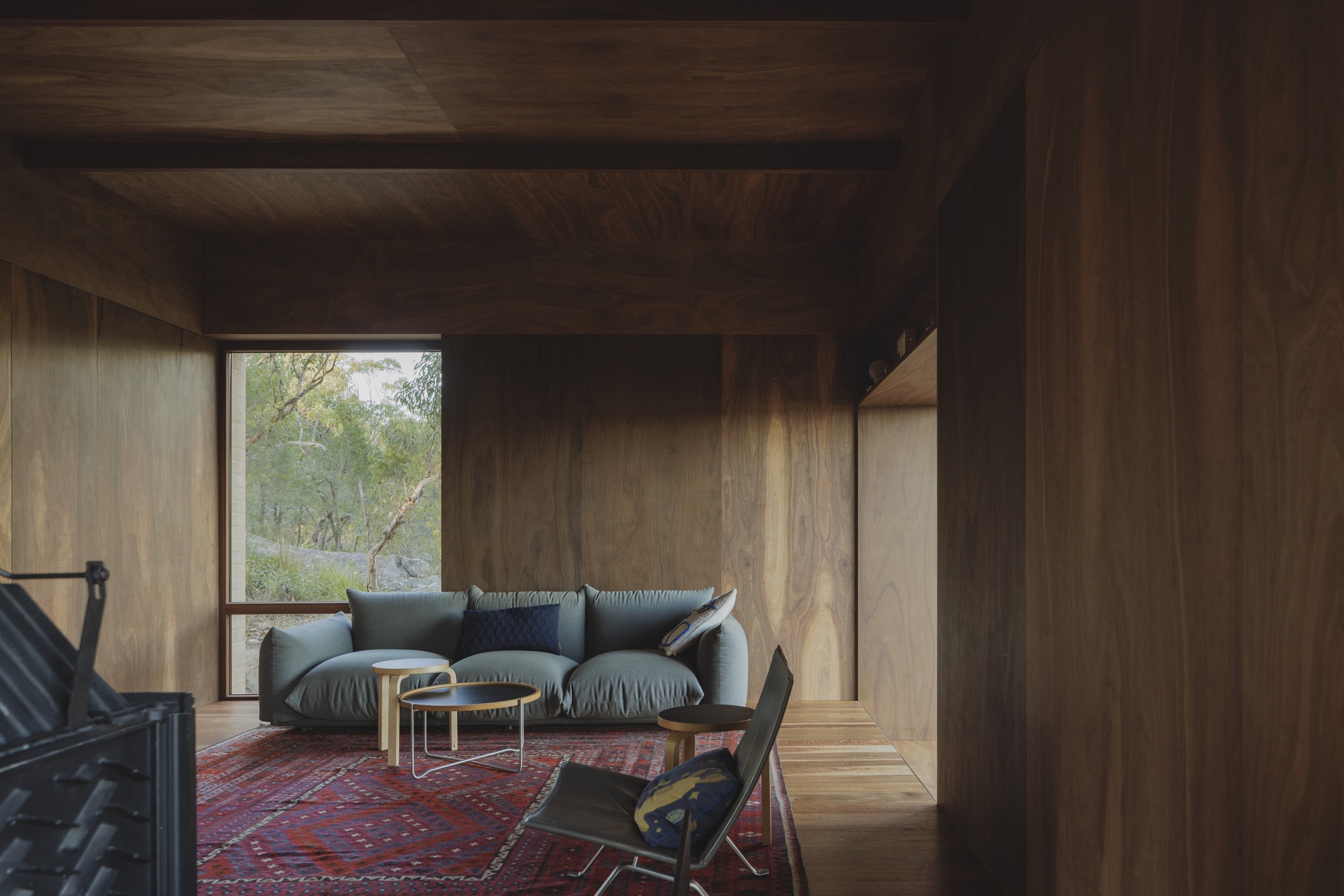 Timber-lined living room with a grey sofa, red patterned rug and a tall window framing bushland in daylight