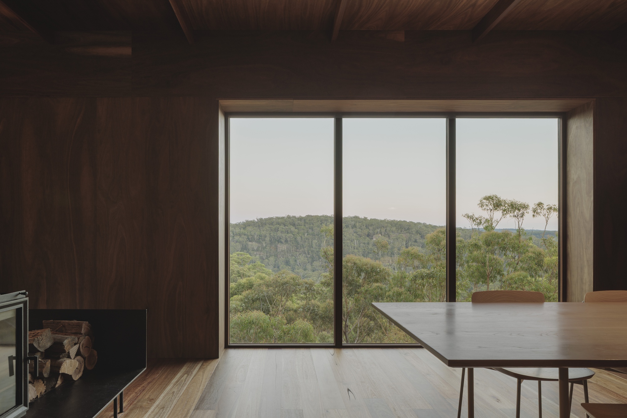 Dining area with a long timber table facing a triple-pane window onto forested hills at dusk, firewood stacked beside the stove