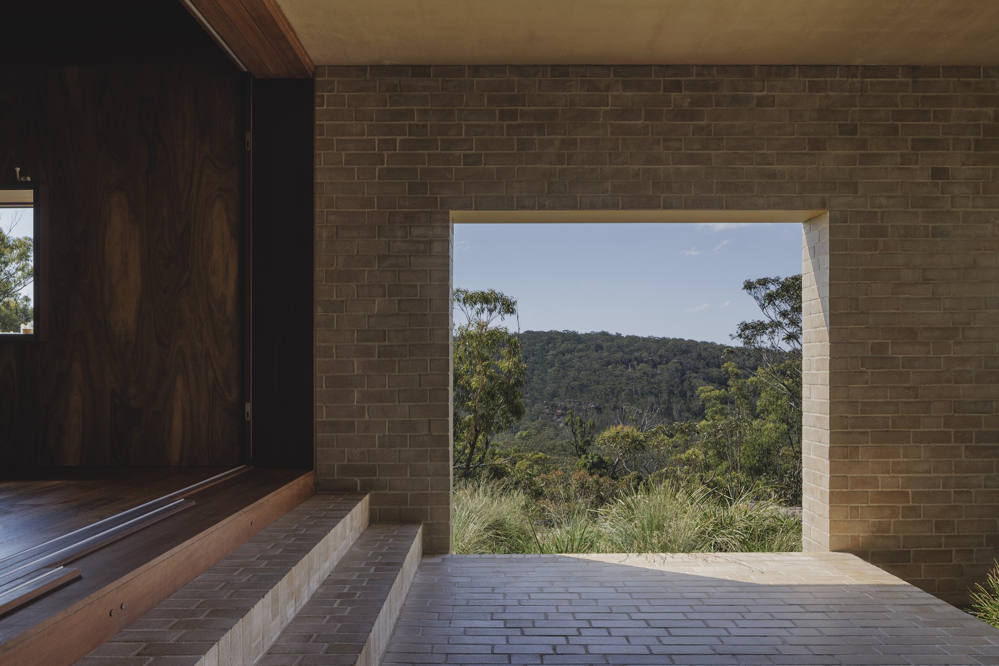 Brick-paved porch with a stepped threshold and a square wall opening framing forested hills and grasses under a blue sky