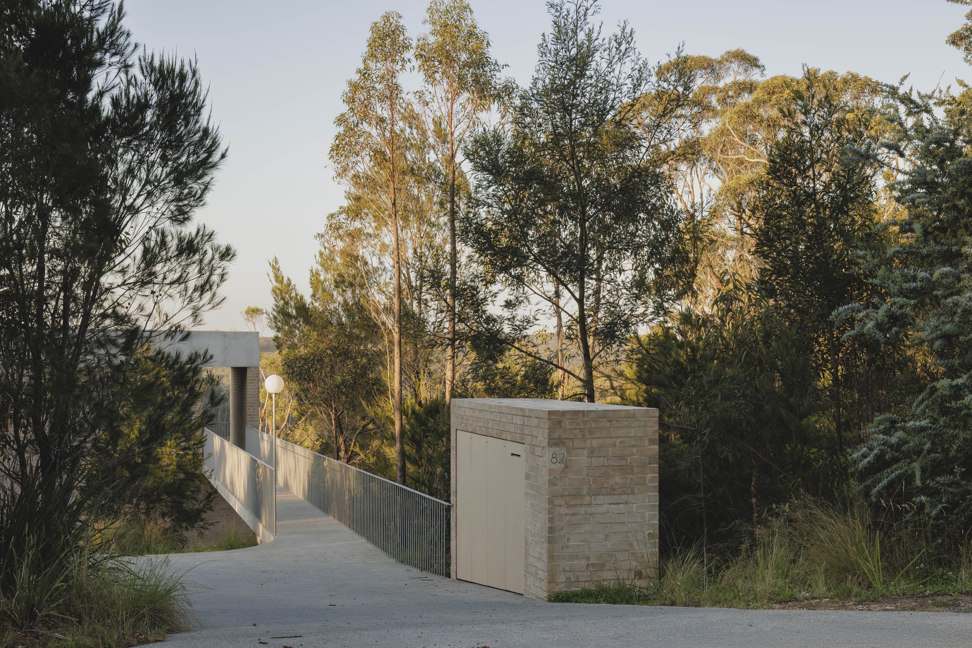 Concrete entry ramp with steel balustrade passing a brick utility enclosure with a timber door, lit by low evening sun