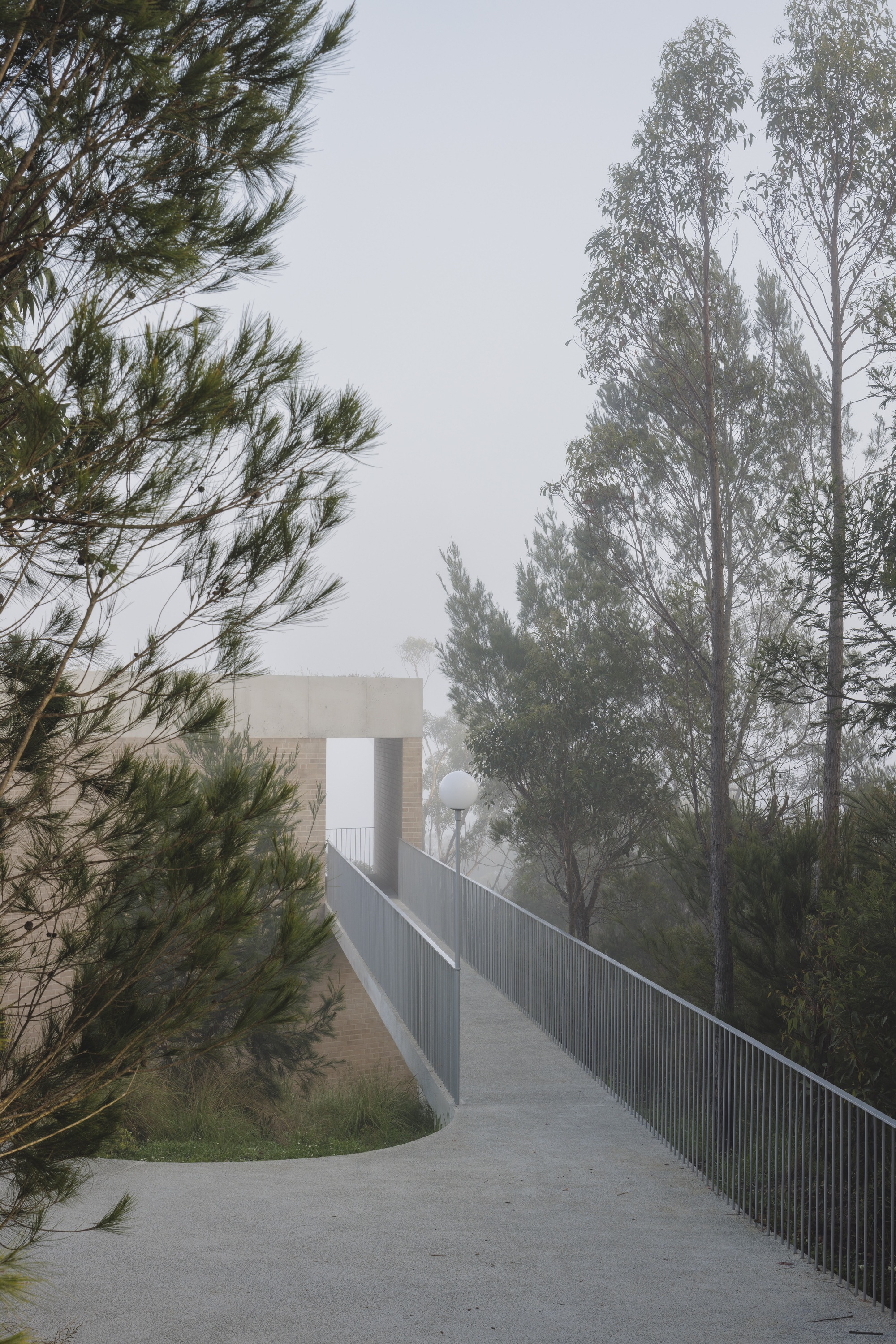 Concrete ramp with slim steel railings and a globe lamp leading toward a brick volume with a concrete lintel in thick fog