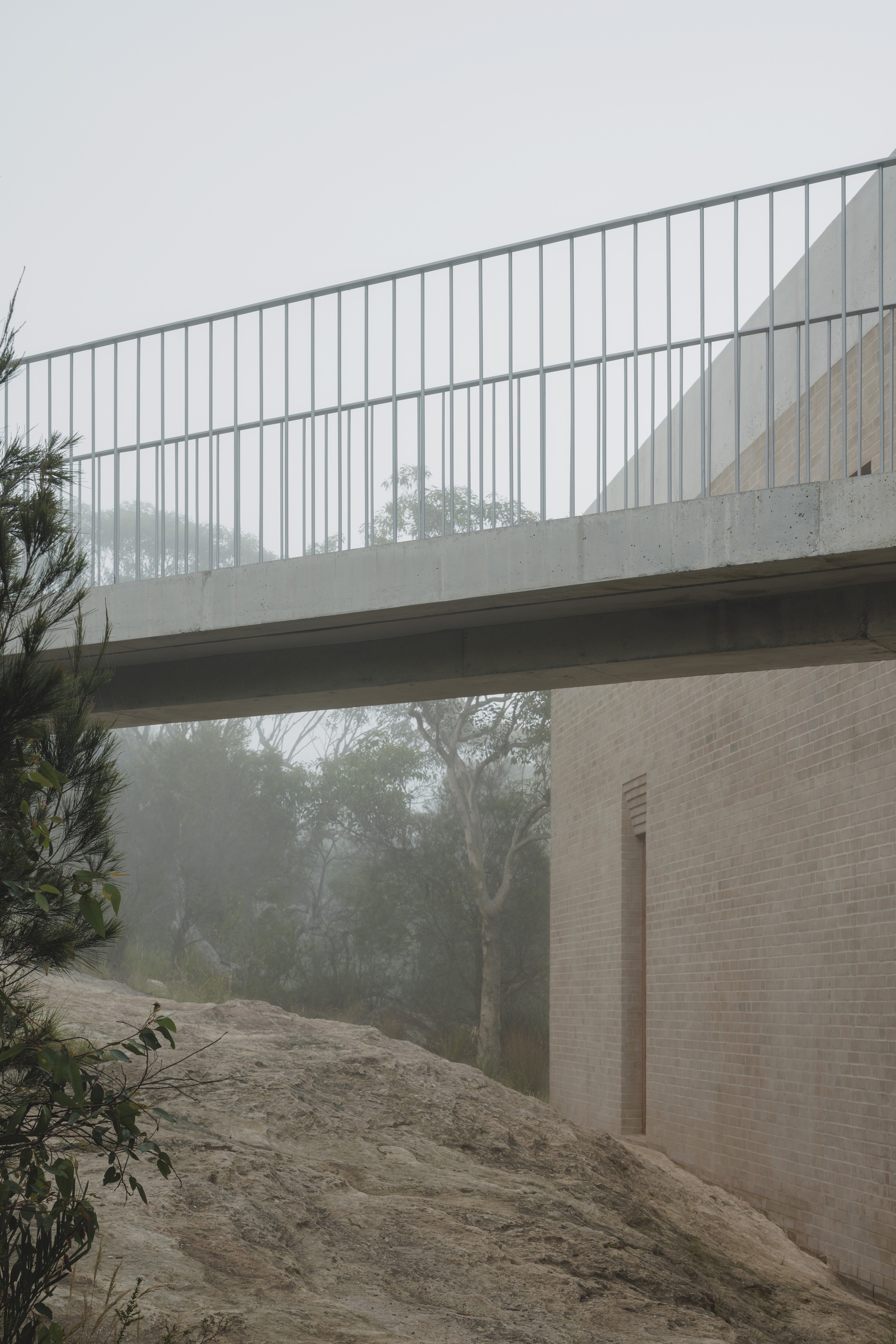 Concrete bridge with a thin steel balustrade crossing above a rocky slope and a brick wall in morning mist