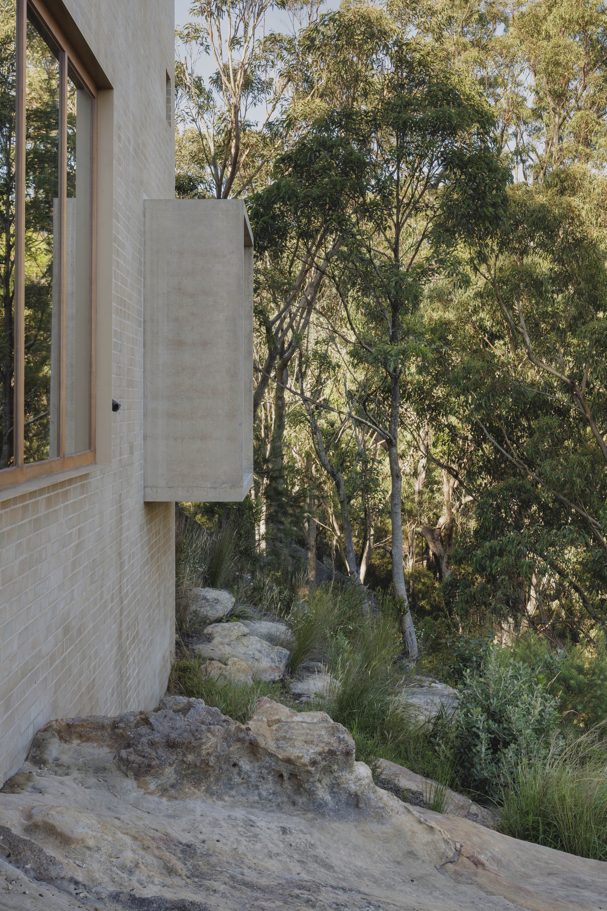 Pale brick wall with a timber-framed window and concrete bay projecting above sandstone boulders and native grasses, with eucalyptus trees behind