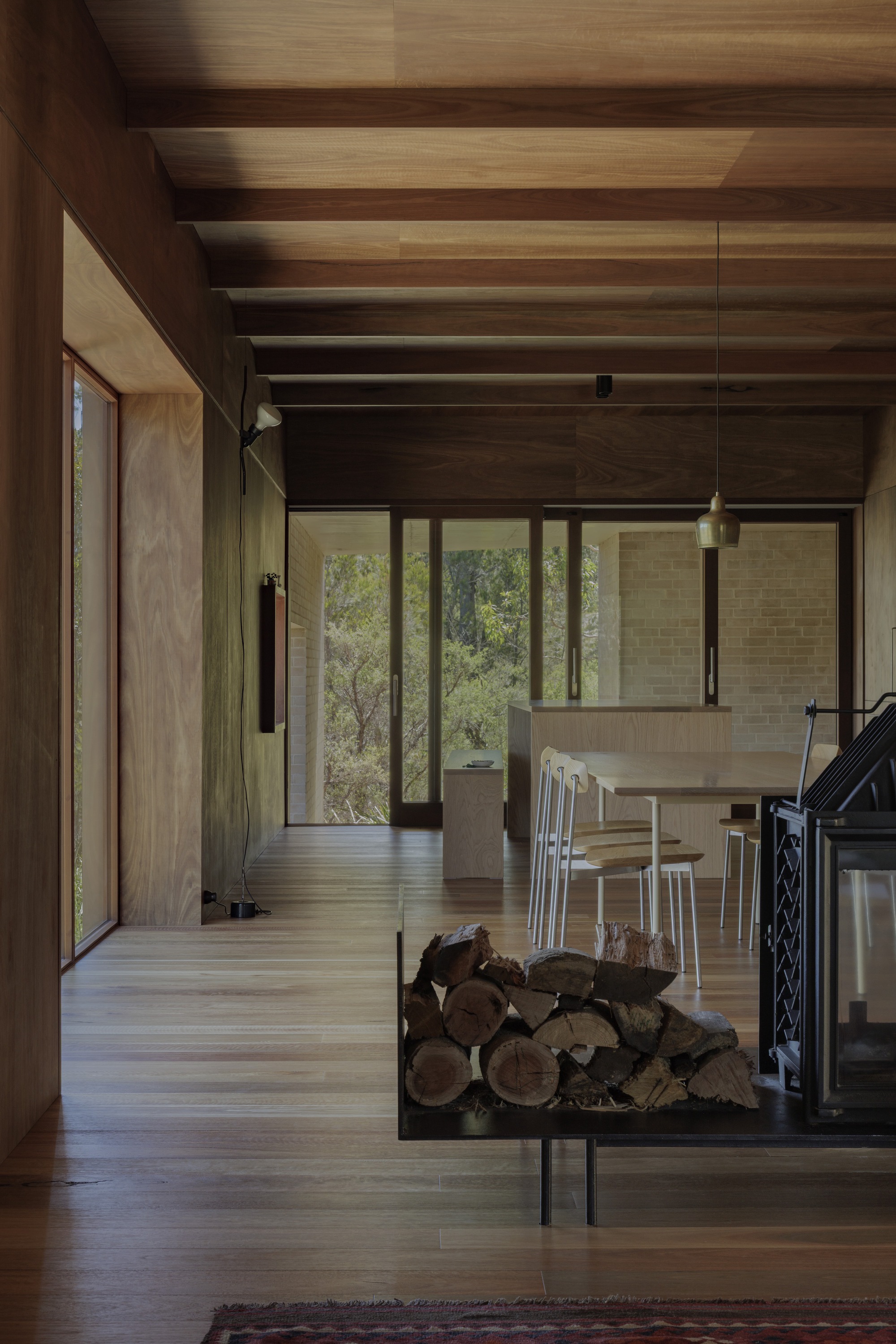 Timber-lined living space with exposed beams, a stacked firewood plinth and a freestanding fireplace, looking toward glazed doors and bushland