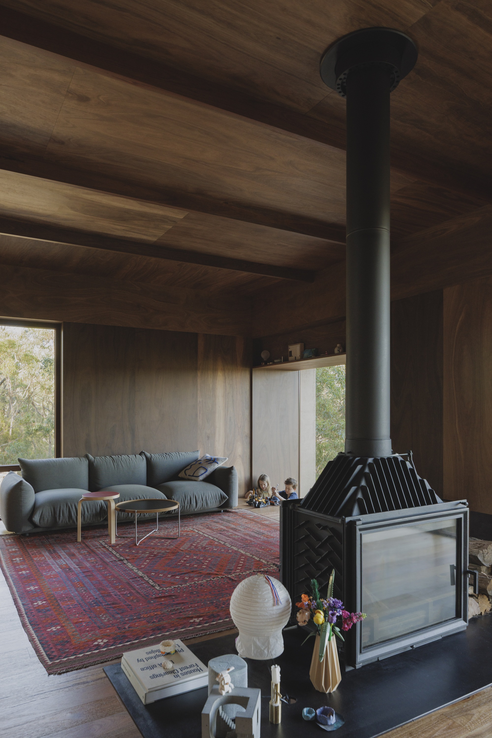 Living room with a grey sofa, red patterned rug and black flue fireplace, with two children playing by a window in the afternoon light